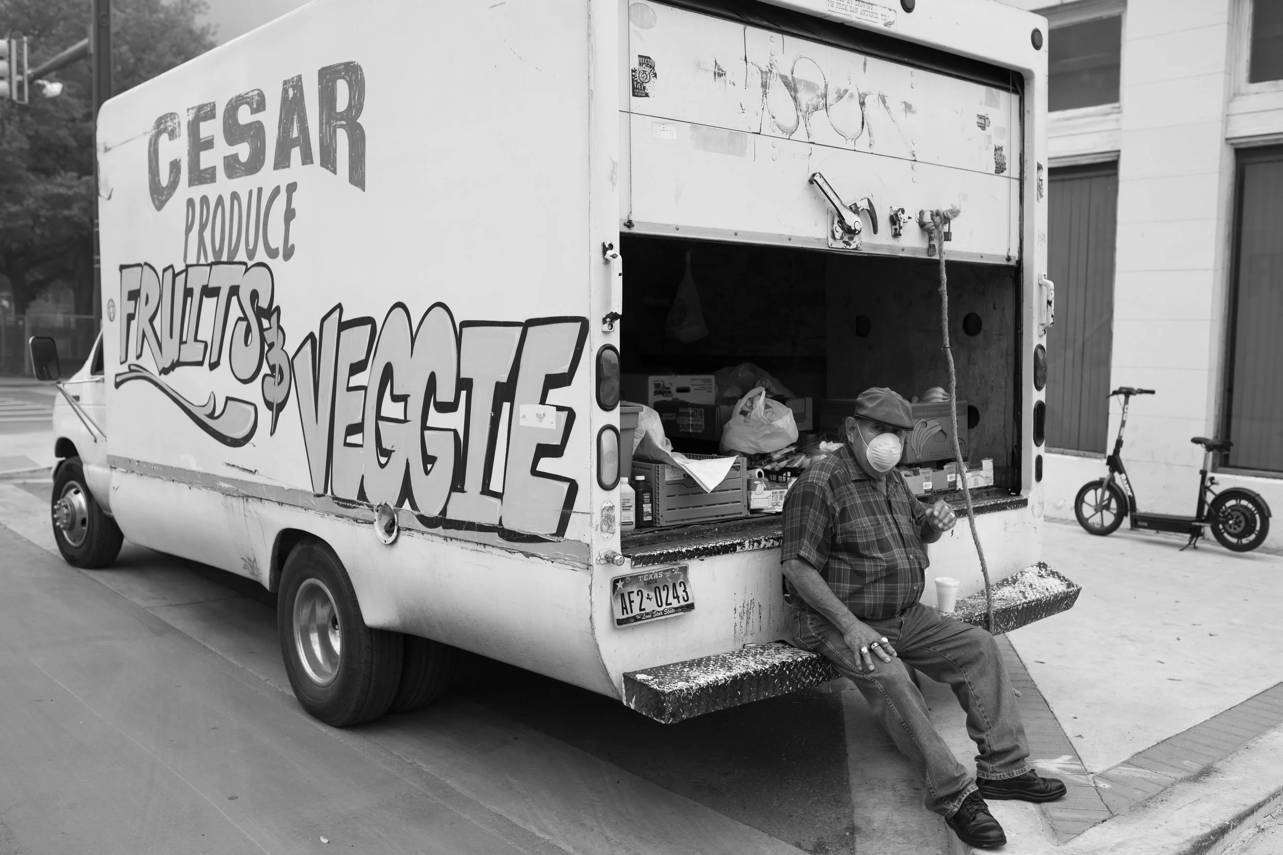 A person wearing a face mask and a hat is sitting on the back step of a truck labeled "Gesar Produce, Fruits & Vegetables". The truck is parked on the side of a city street, with a bicycle and a building visible in the background.