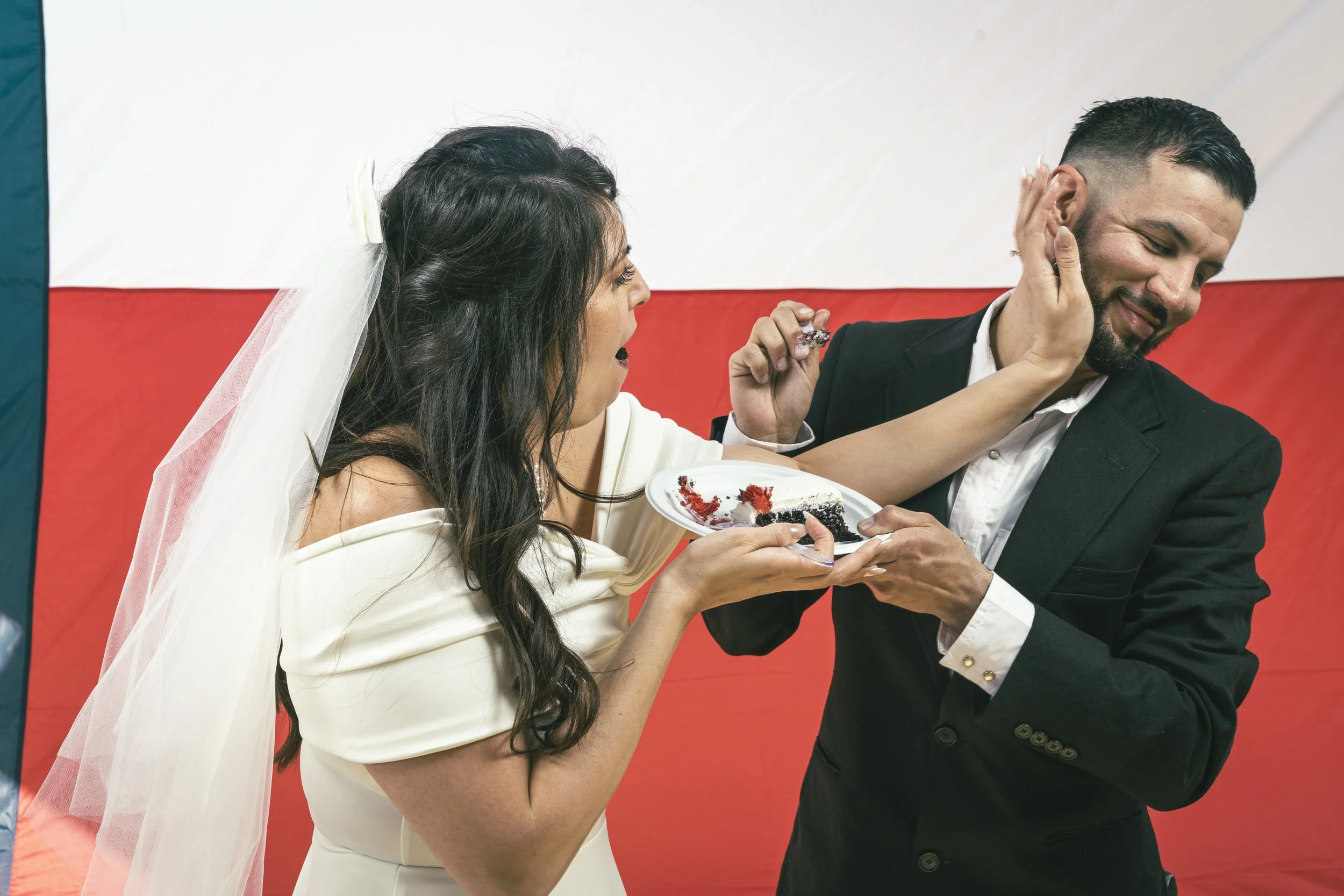 A woman in a white dress and wedding veil playfully smears cake on a man in a black suit. They are at a celebration with a red, white, and green background.