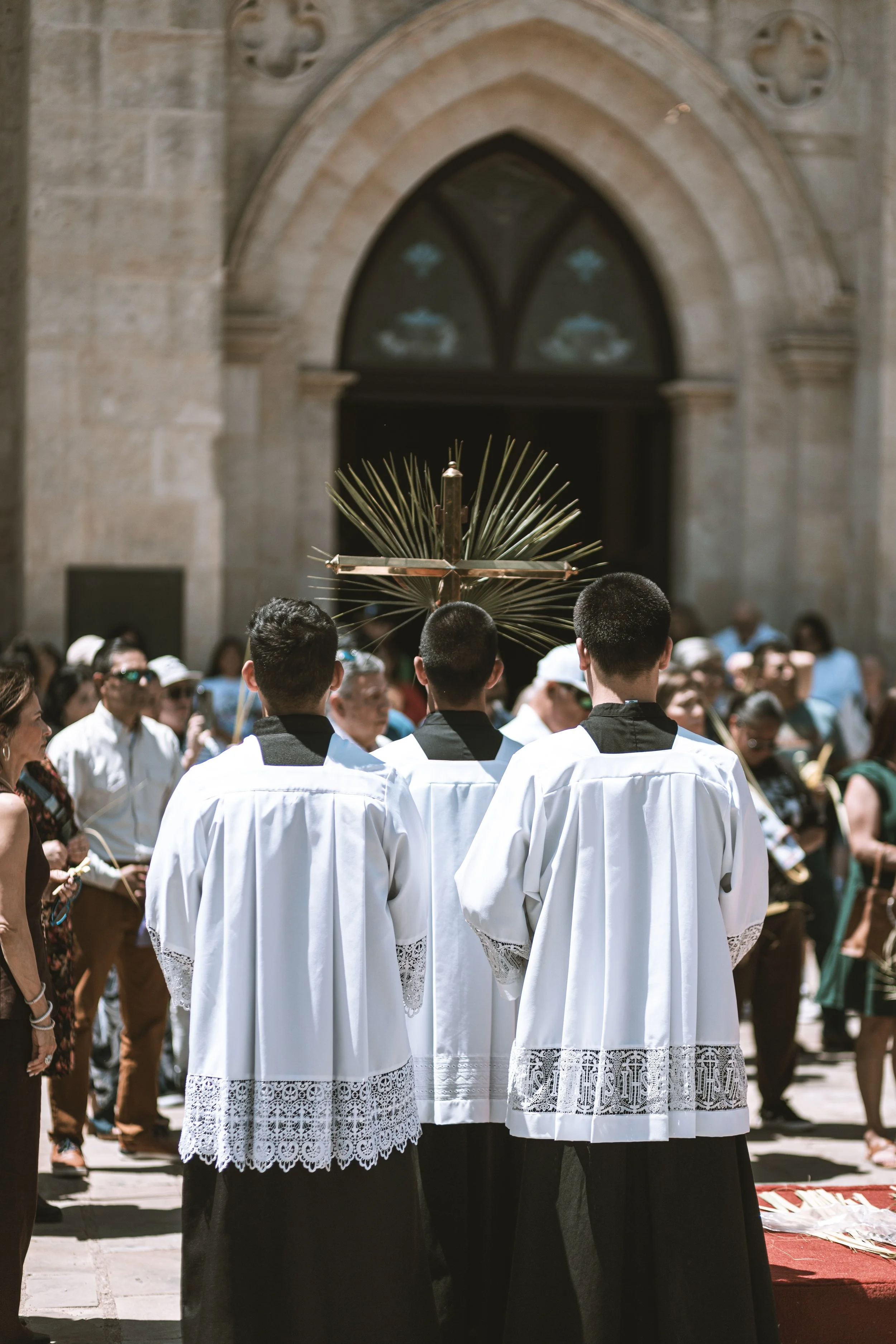 People in religious robes participating in a Catholic procession in front of a church with an arched stained glass window, carrying a religious flower cross.