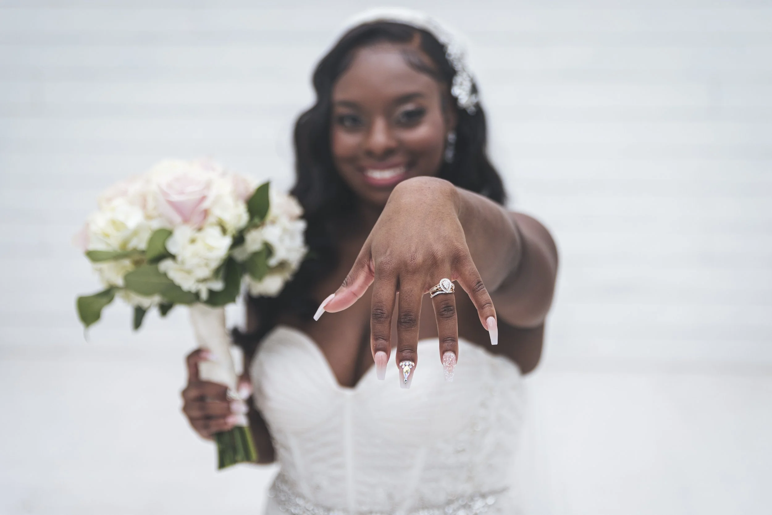 A woman in a white dress shows her hand with an engagement ring, smiling and holding a bouquet of white and pink flowers.