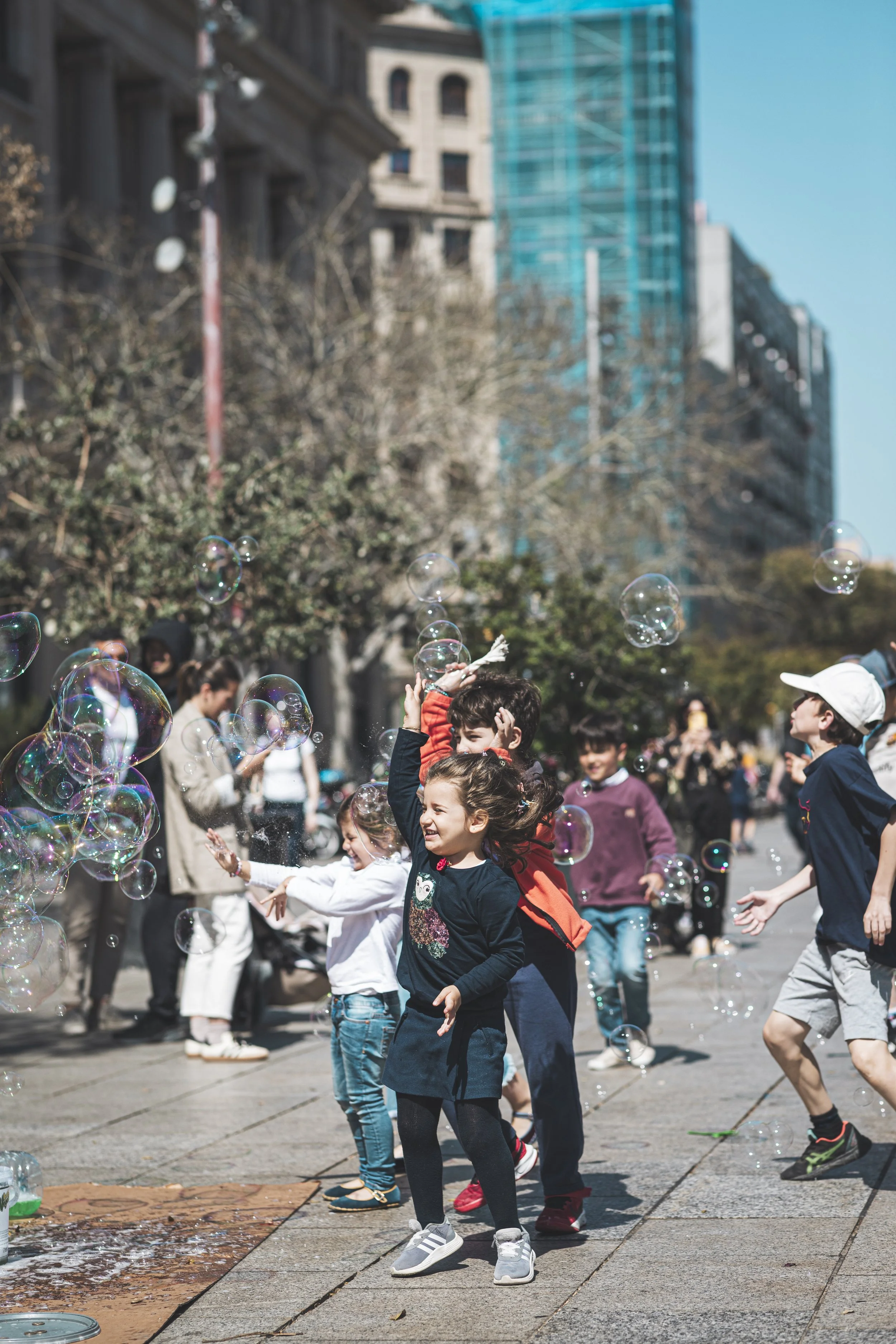 Children playing and chasing bubbles in an outdoor urban space during daytime, with buildings and trees in the background.