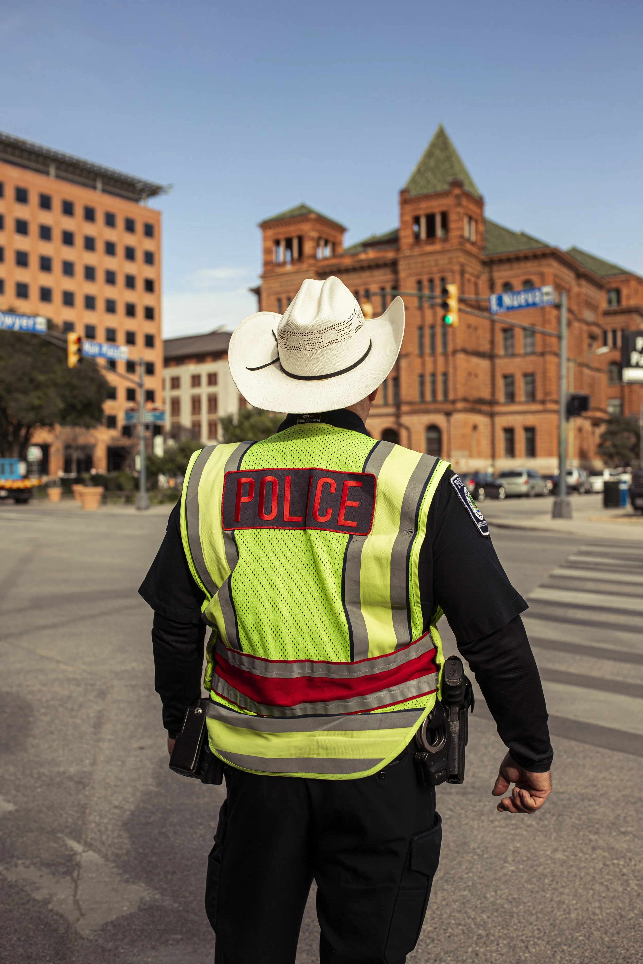 Back of a police officer wearing a yellow reflective vest and a white cowboy hat, standing in the middle of a street with historic brick buildings in the background.