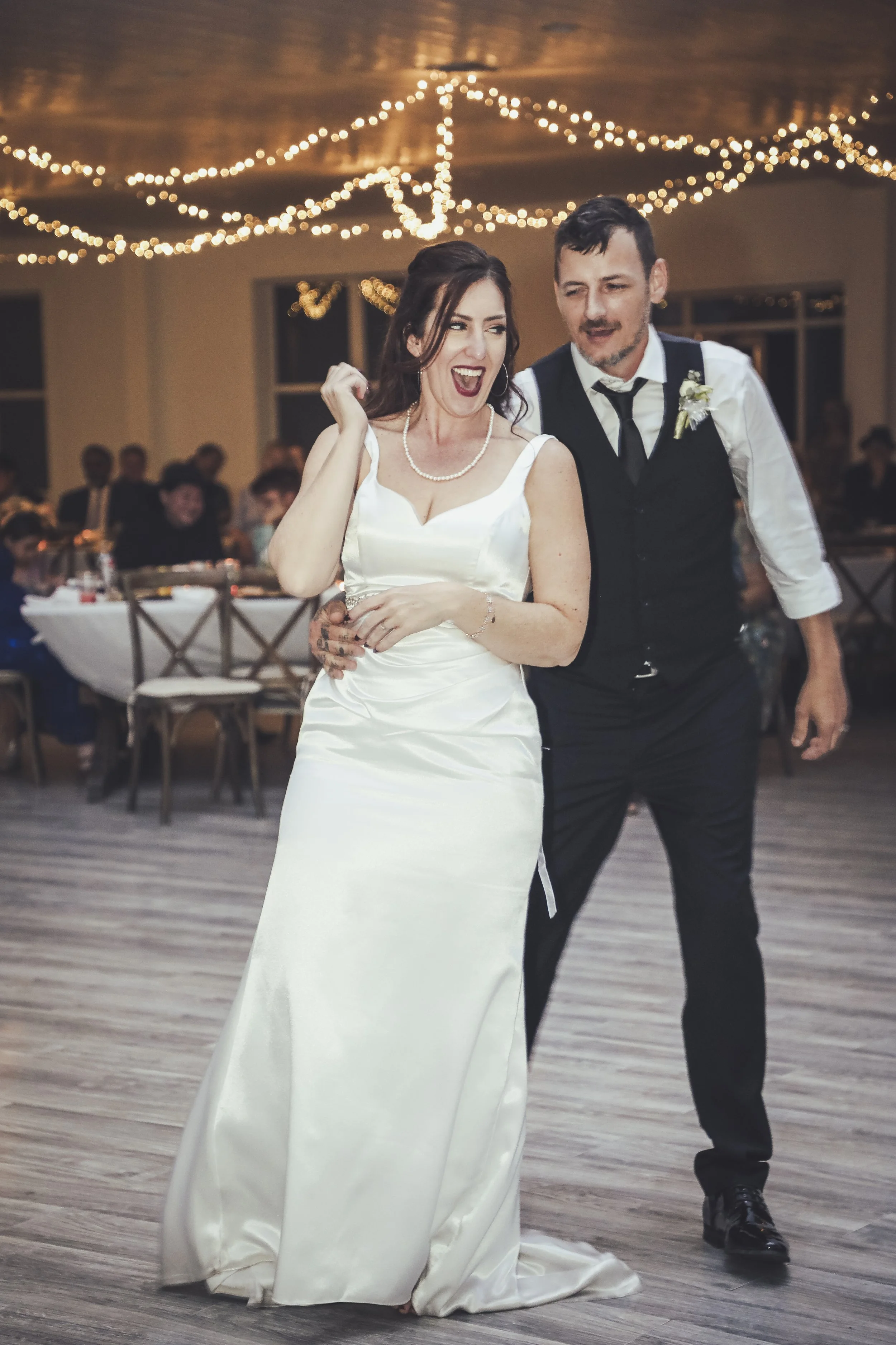 A bride and groom dancing at their wedding reception, with the bride wearing a white wedding gown and the groom in a black vest with a white shirt and black tie, under string lights.