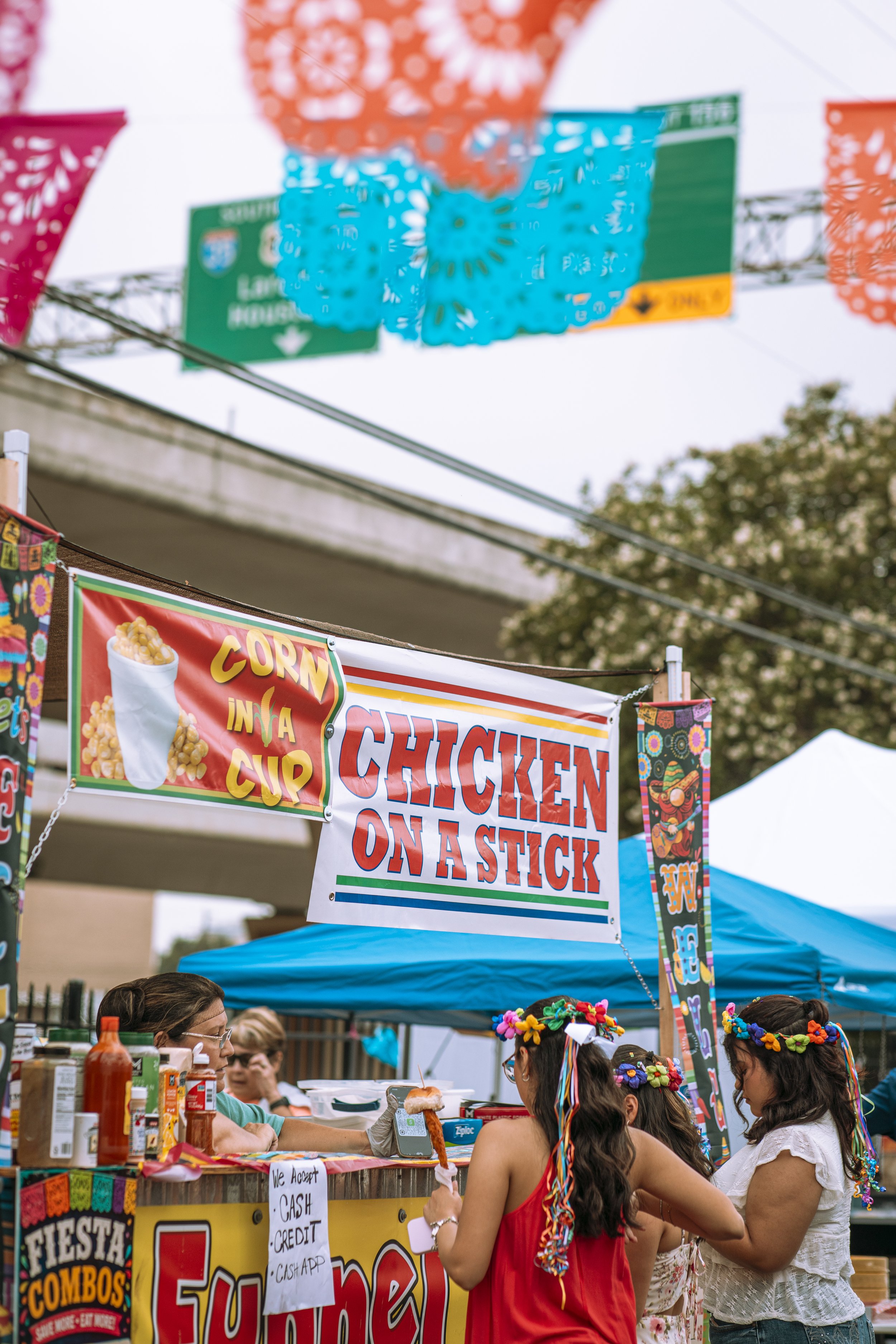 People at a food stand selling fried chicken on a stick, with colorful banners and decorations at an outdoor festival.