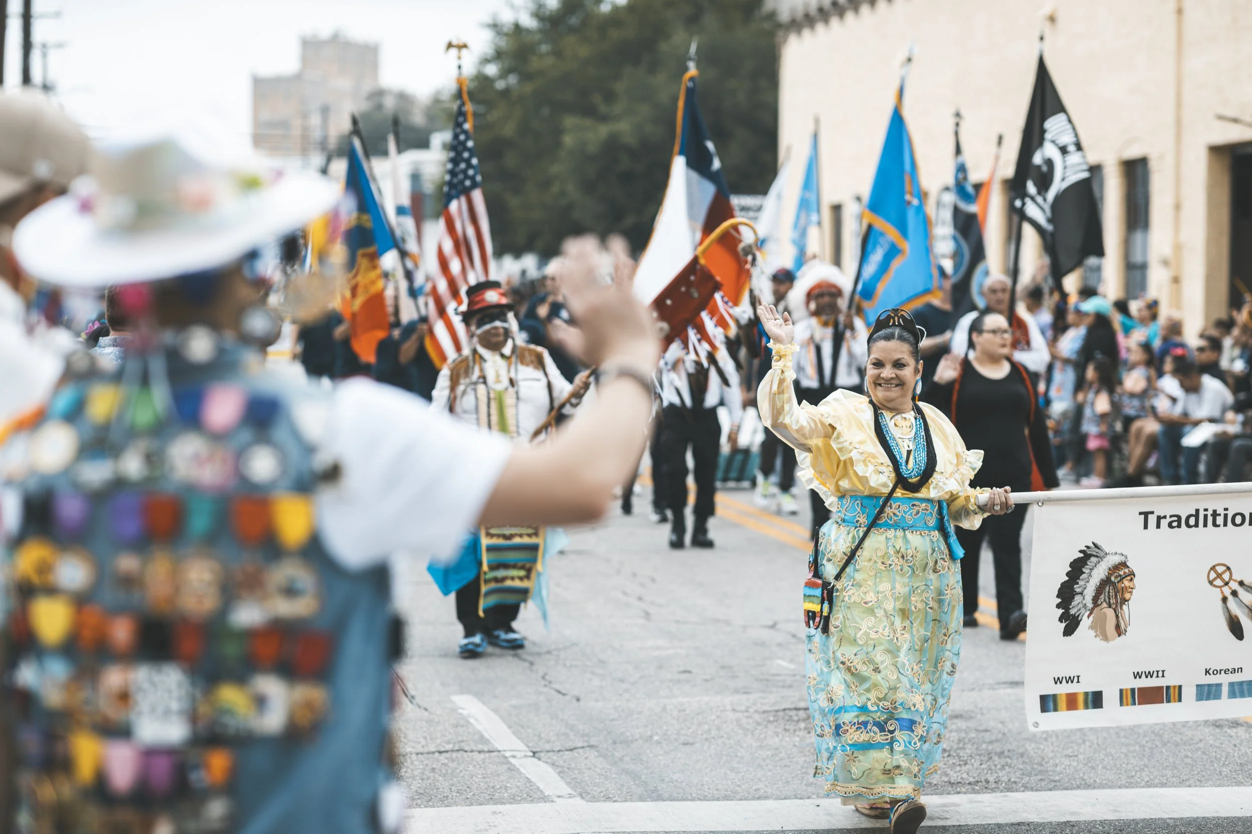 A woman wearing a yellow and blue native dress and jewelry waves in a parade with flags in the background, alongside participants in rainbow-colored costumes and other parade attendees.
