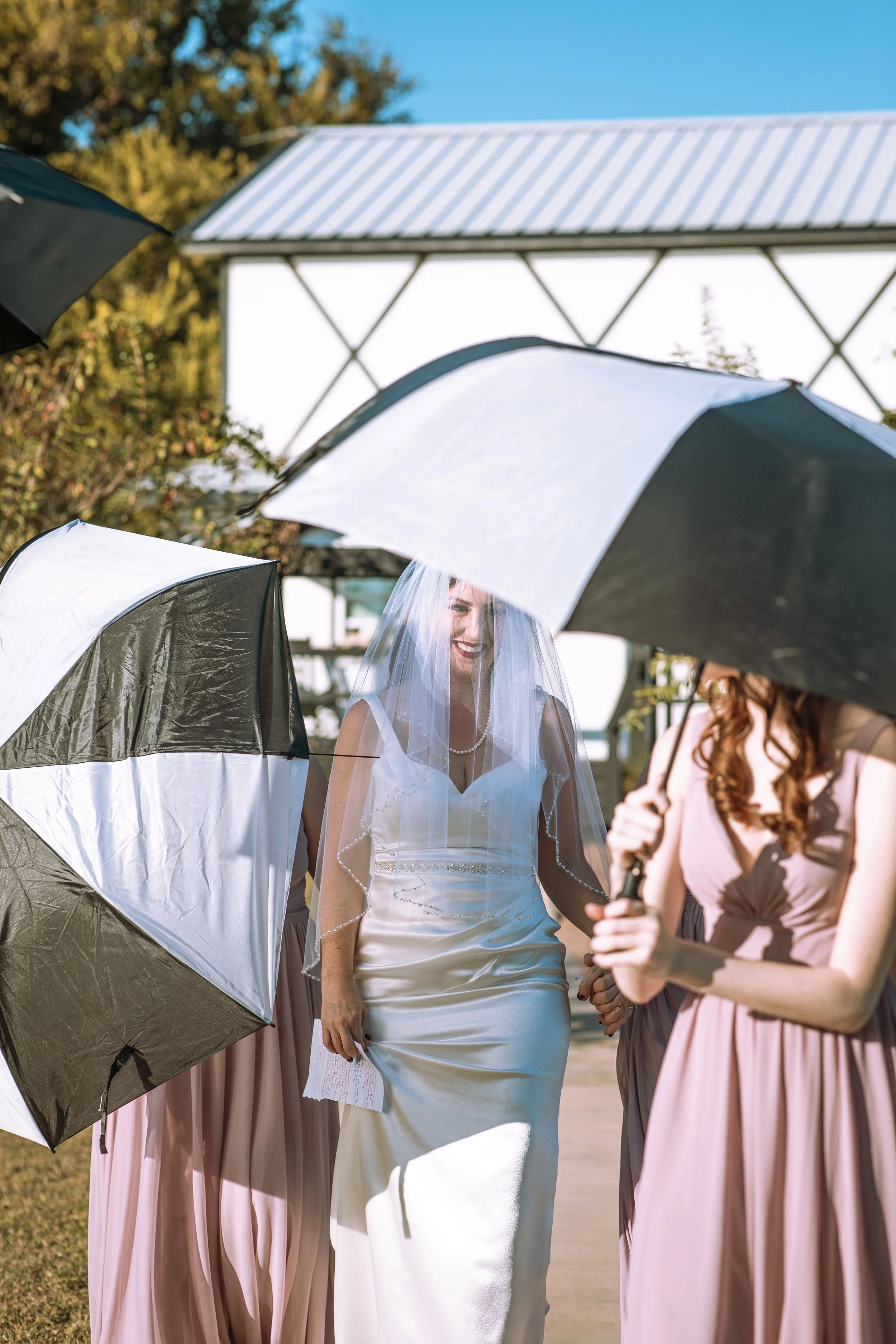 A bride in a white wedding dress with a veil walking outside, surrounded by bridesmaids in long pink dresses, holding umbrellas on a sunny day.
