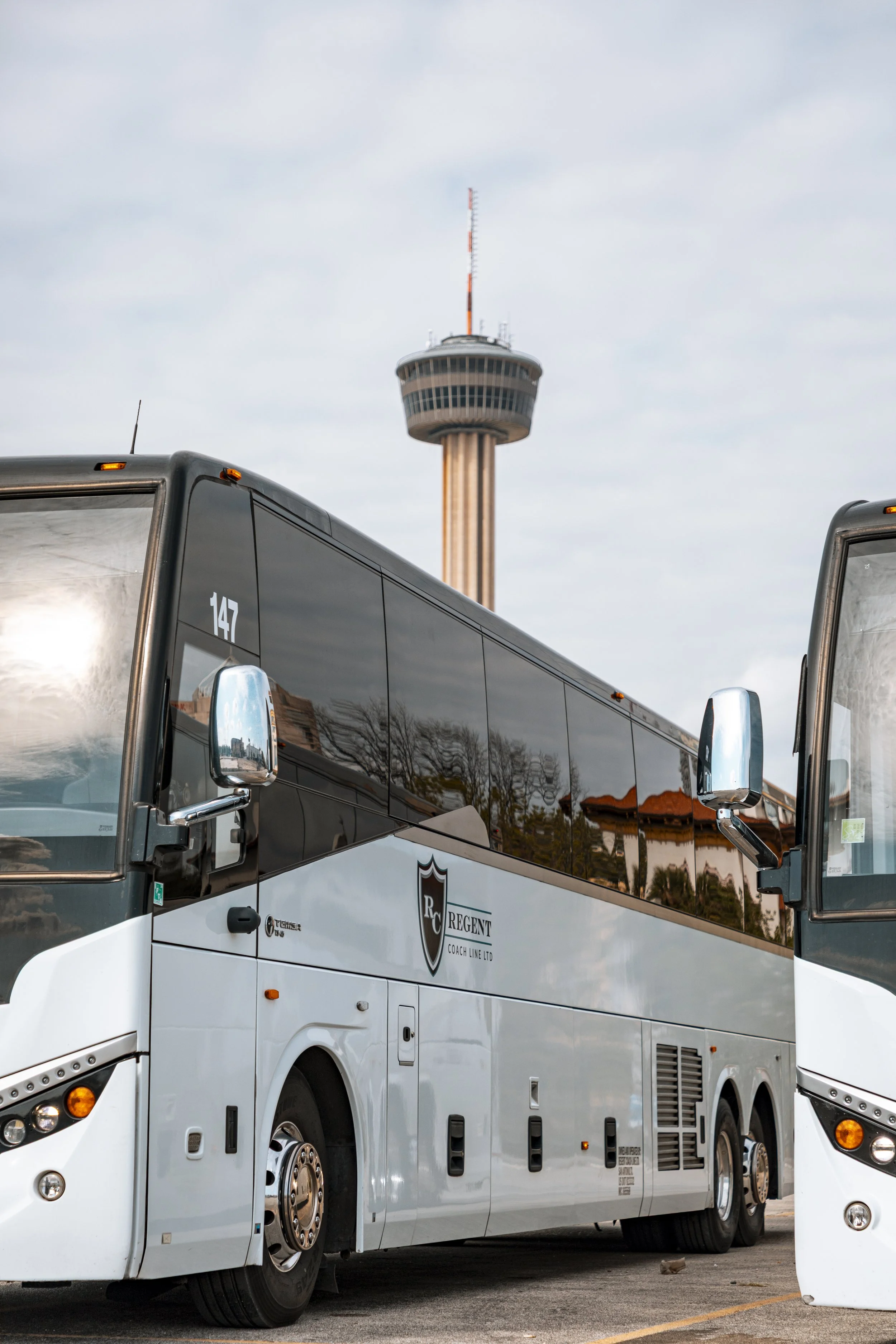 A white luxury bus with the logo 'RC Regent Coach Line Ltd' parked in front of a tall observation tower. The bus has the number 147 and mirrors reflecting part of the environment. The background shows a cloudy sky.