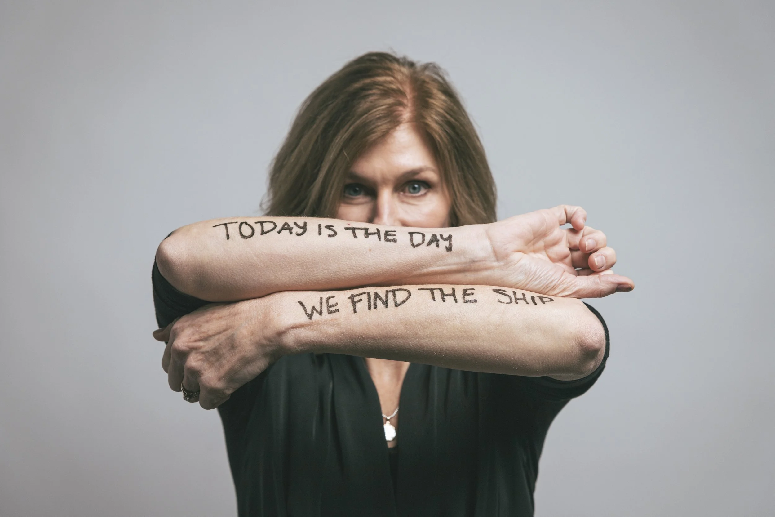 A woman with brown hair and blue eyes is holding her arms crossed in front of her face. The inside of her arms has handwritten text that reads, "TODAY IS THE DAY" and "WE FIND THE SHIP."