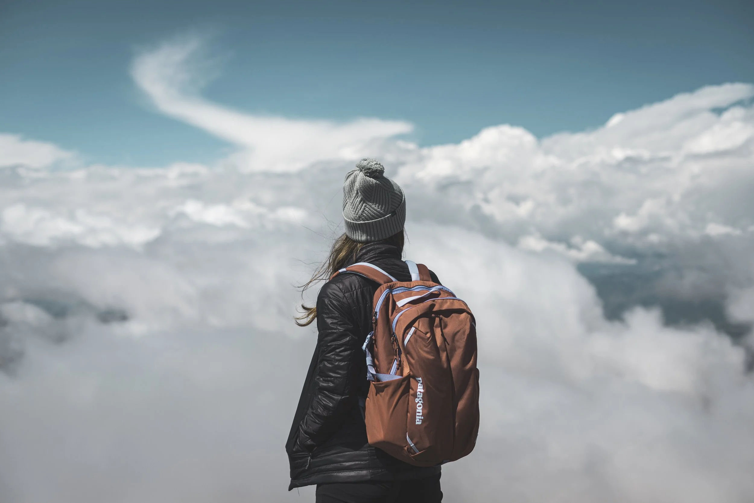 A person with long hair, wearing a gray knitted hat, black jacket, and carrying a brown backpack, standing outdoors and looking at the cloudy sky.