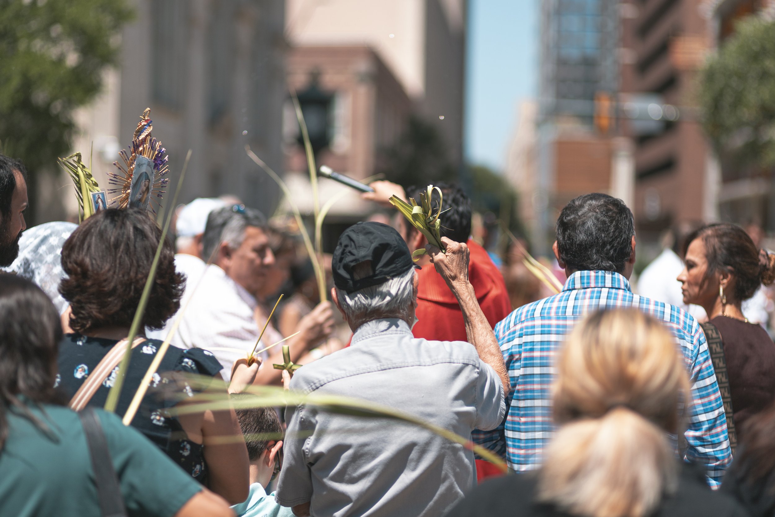 A crowd of people gathered in an outdoor urban setting, holding small palm fronds and religious images during a procession or celebration.
