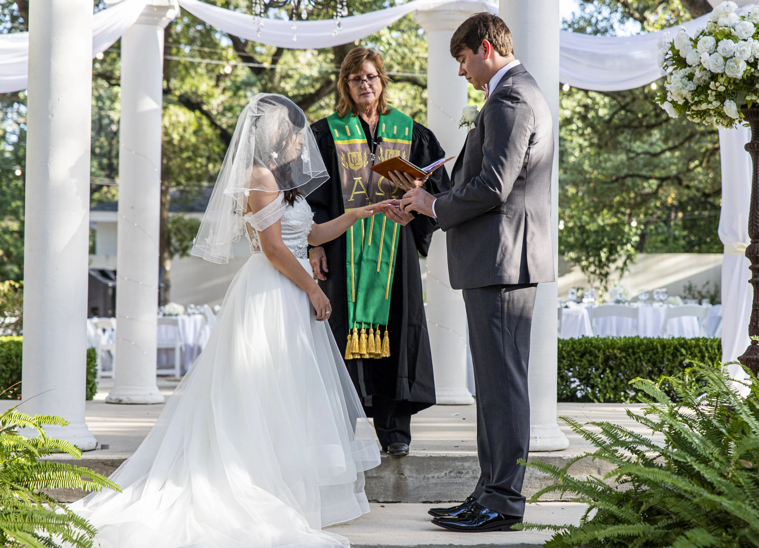 A couple performing a wedding ceremony outdoors under a white draped canopy with a floral arrangement on the side. The bride is wearing a white wedding gown with a veil, and the groom is in a gray suit with a white shirt and a boutonniere. An officia