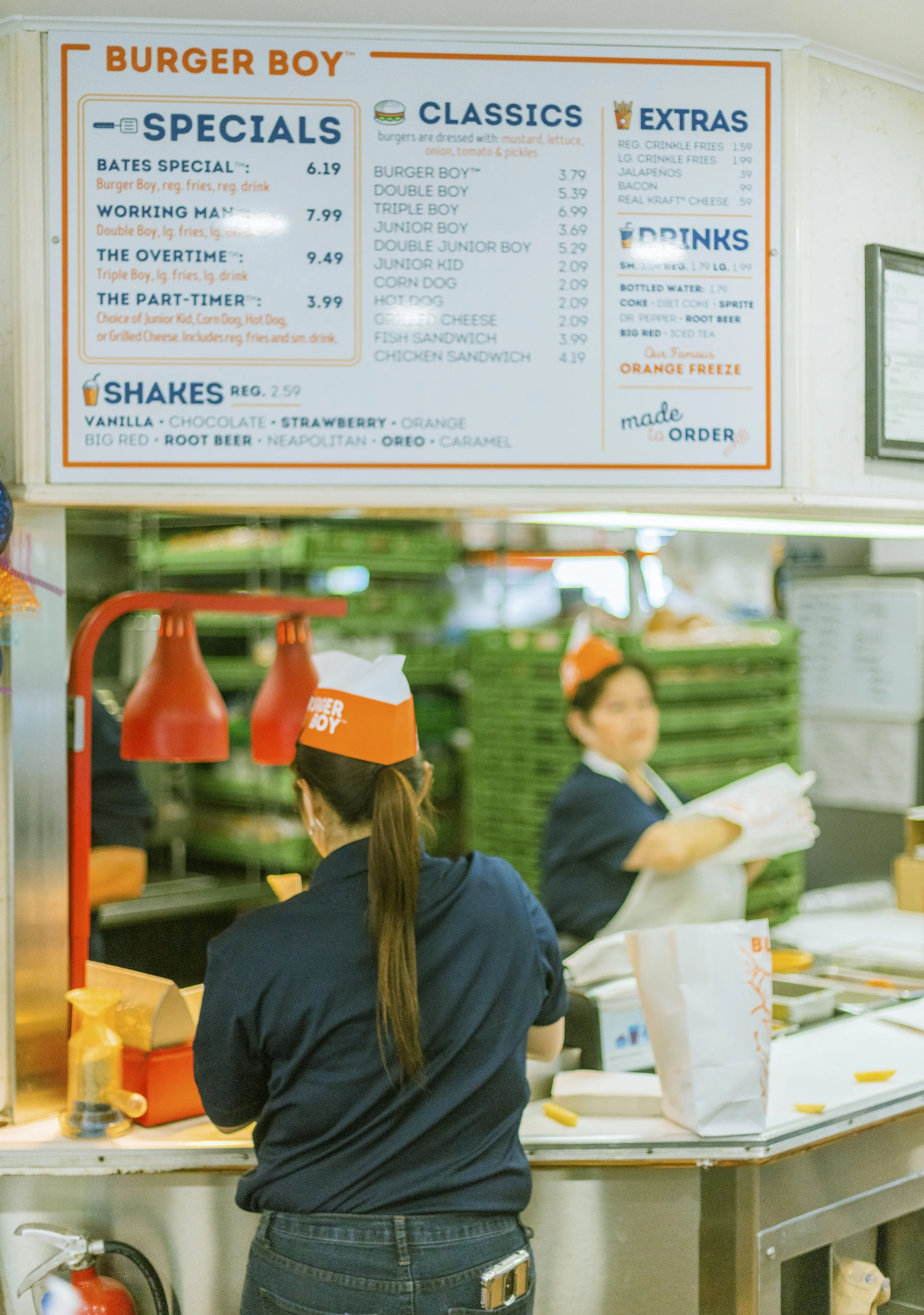 A menu board at Burger Boy featuring specials, classics, extras, and drinks, with employees preparing orders in the kitchen area visible below.