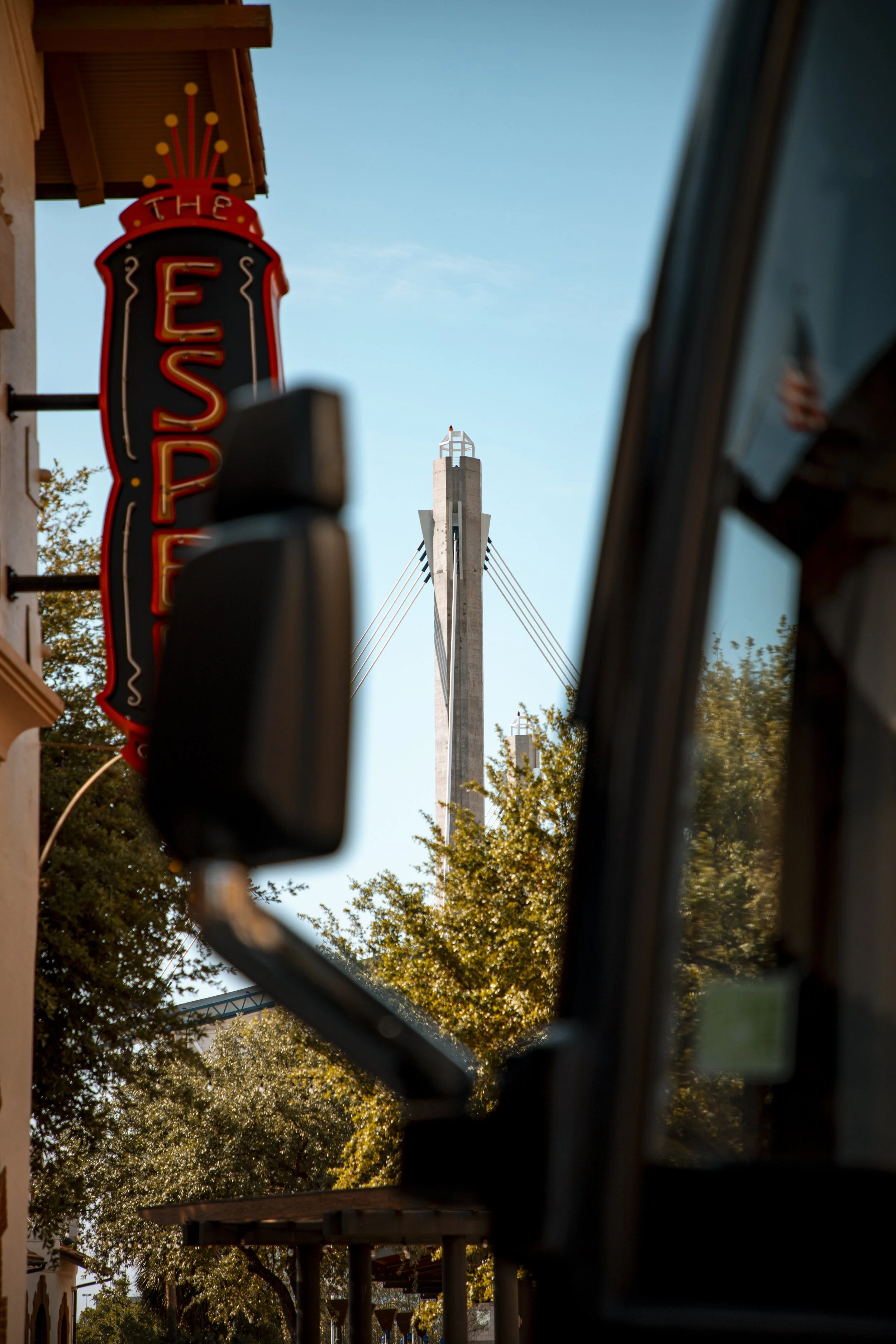 A lit-up neon sign for The Edison, a restaurant or bar, on a building on a street with trees and a bridge in the background, viewed through a car window.