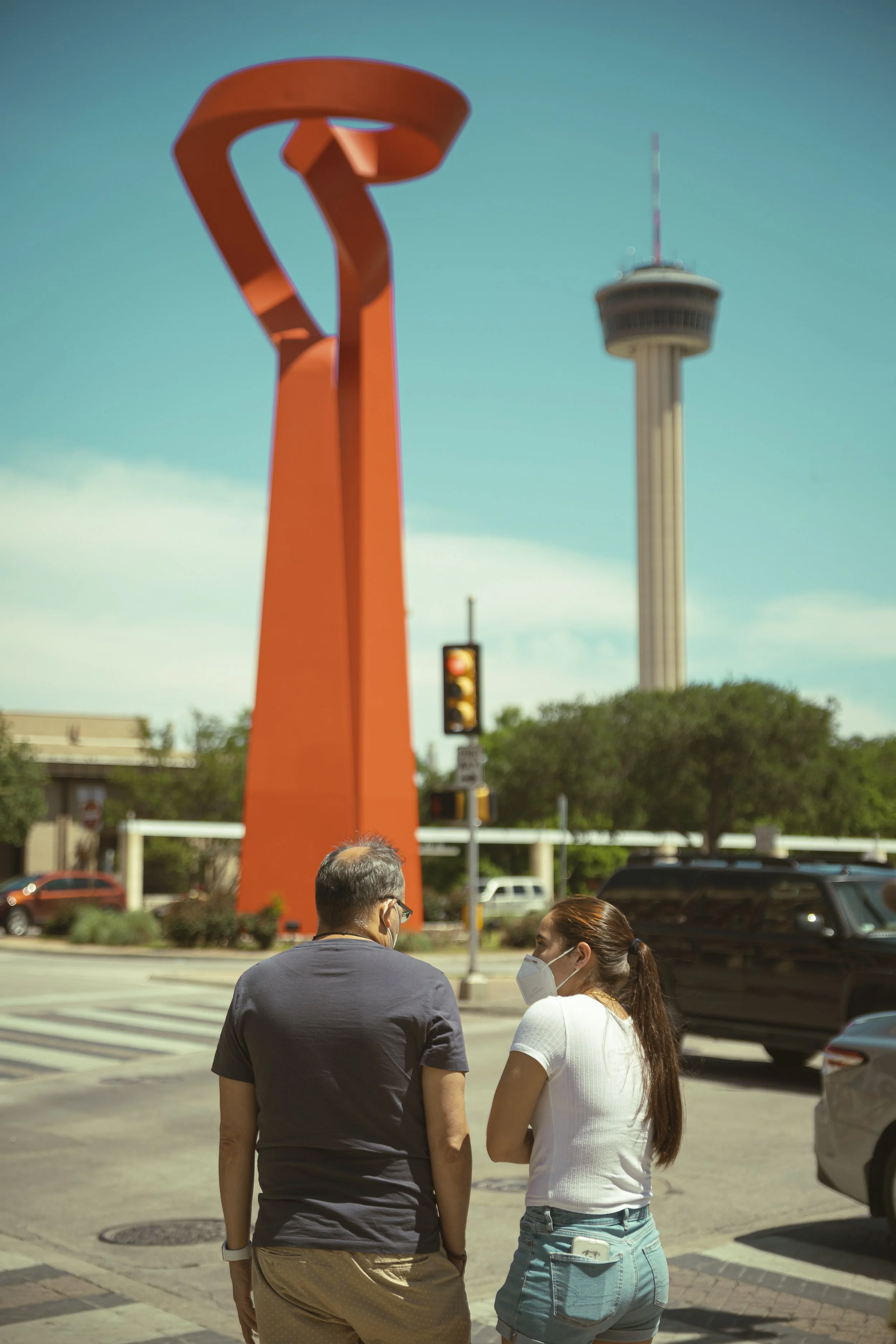 People wearing face masks standing and talking at a crosswalk in an urban area with a tall observation tower and a modern sculpture in the background.