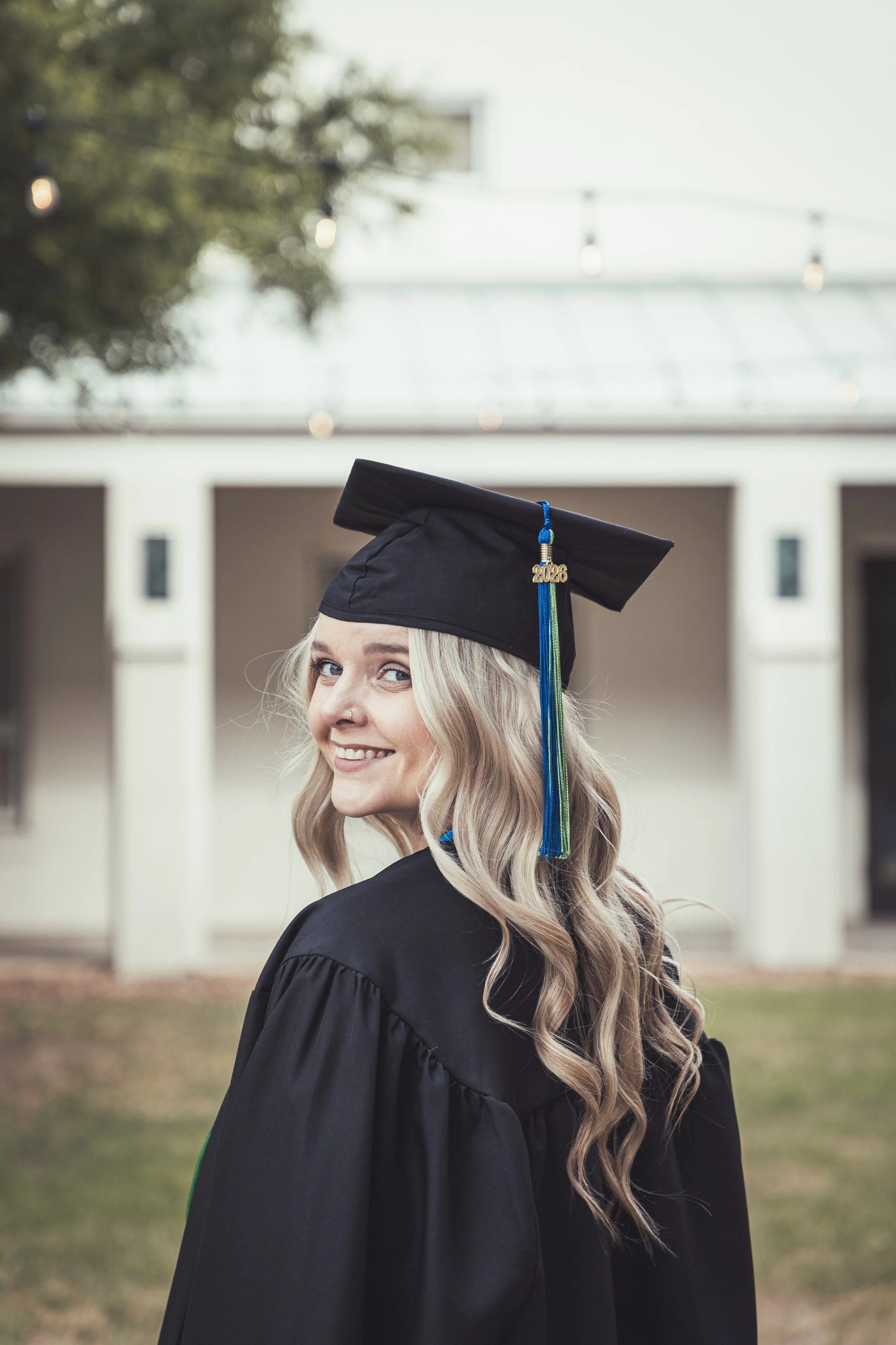 A young woman with long blonde hair, smiling, wearing a black graduation cap with a year "2026" charm and a black gown, outdoors with a building and string lights in the background.