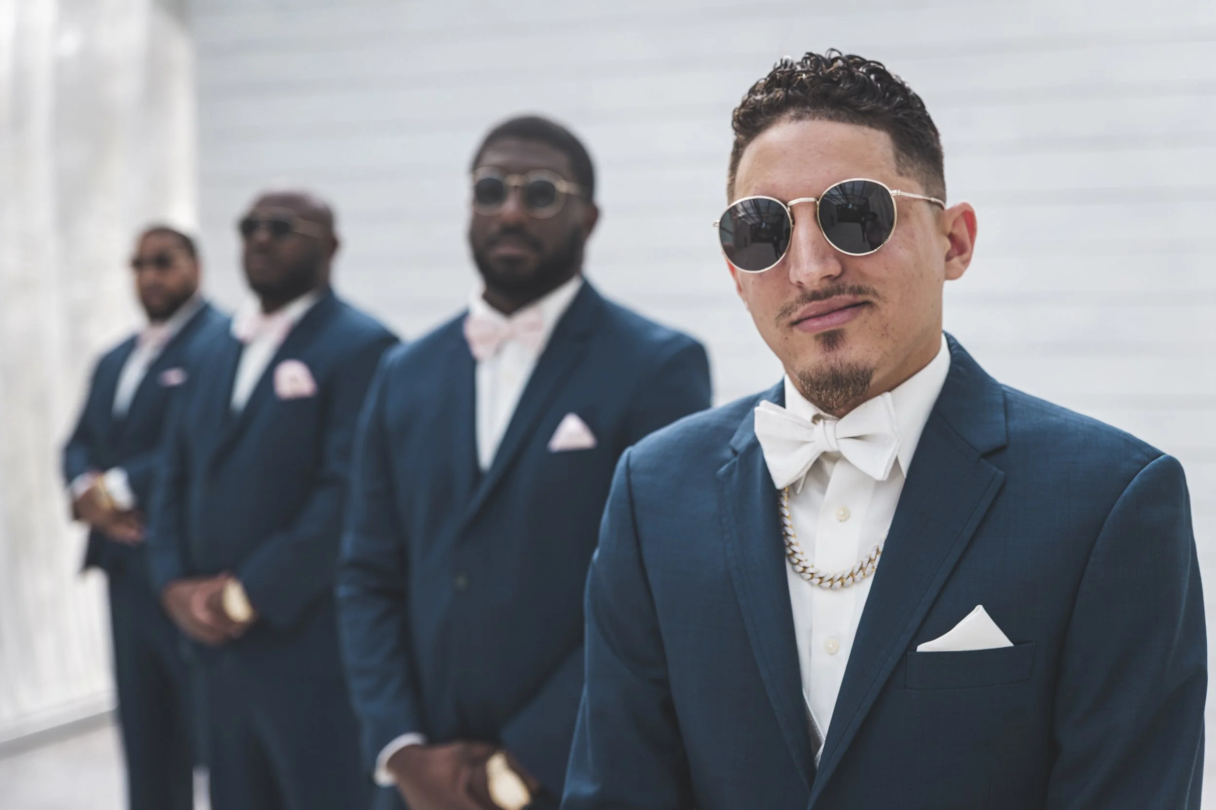 Four stylish men dressed in formal suits with bow ties, wearing sunglasses, standing in a modern, minimalist indoor space.