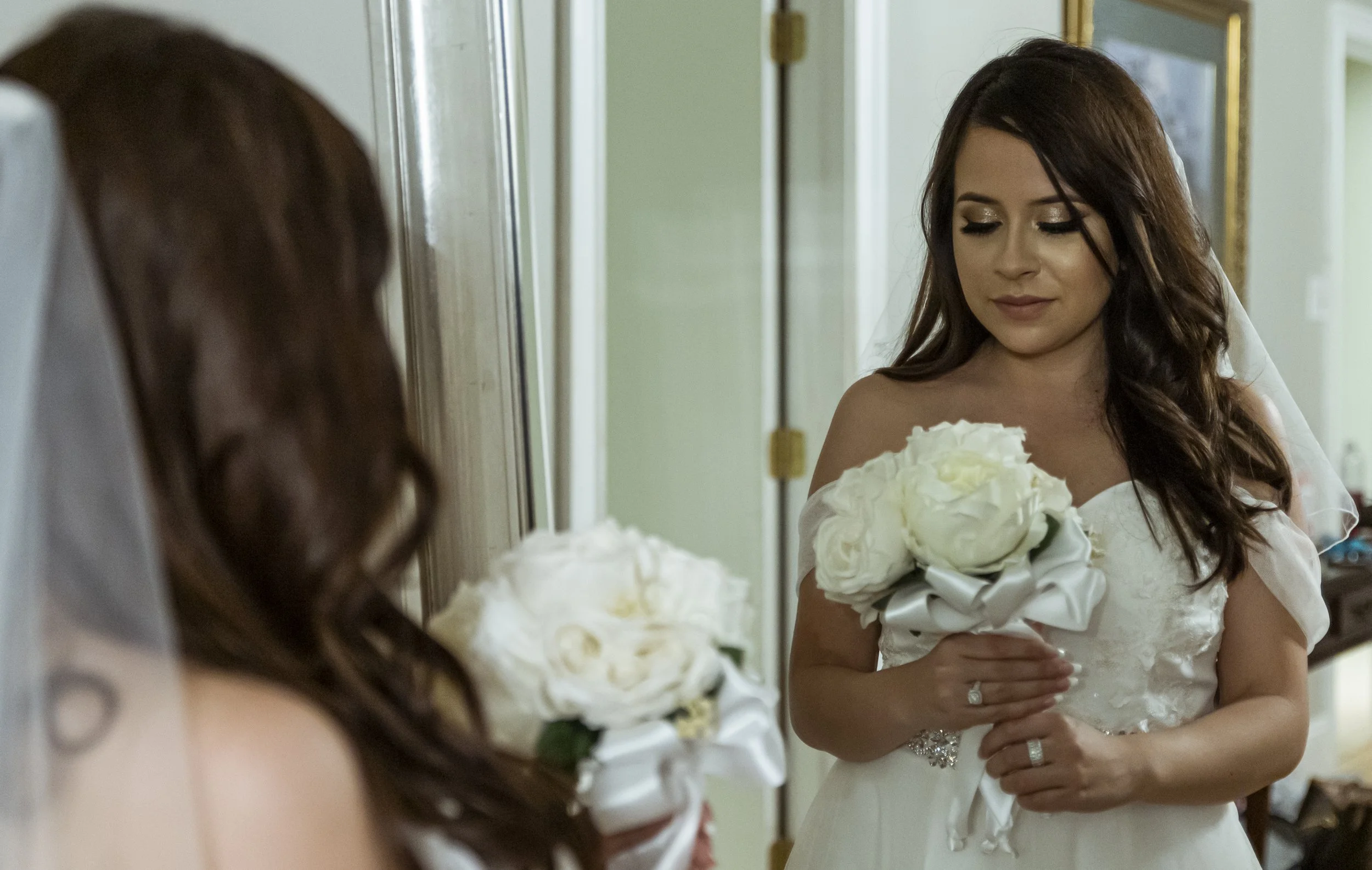 Bride in a white wedding dress holding a bouquet of white roses, looking at her reflection in a mirror.