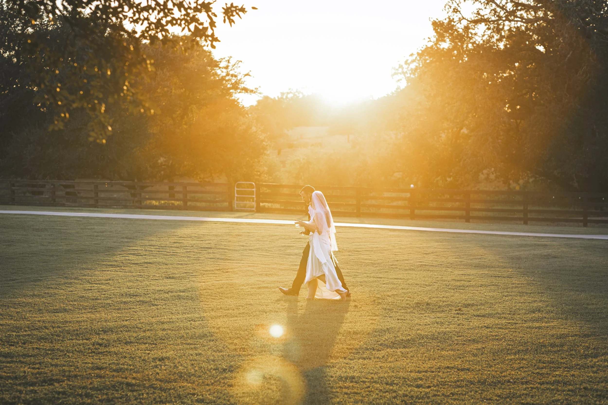A bride and groom walking together on a grassy field during sunset, with the sun shining brightly in the background and trees on either side.