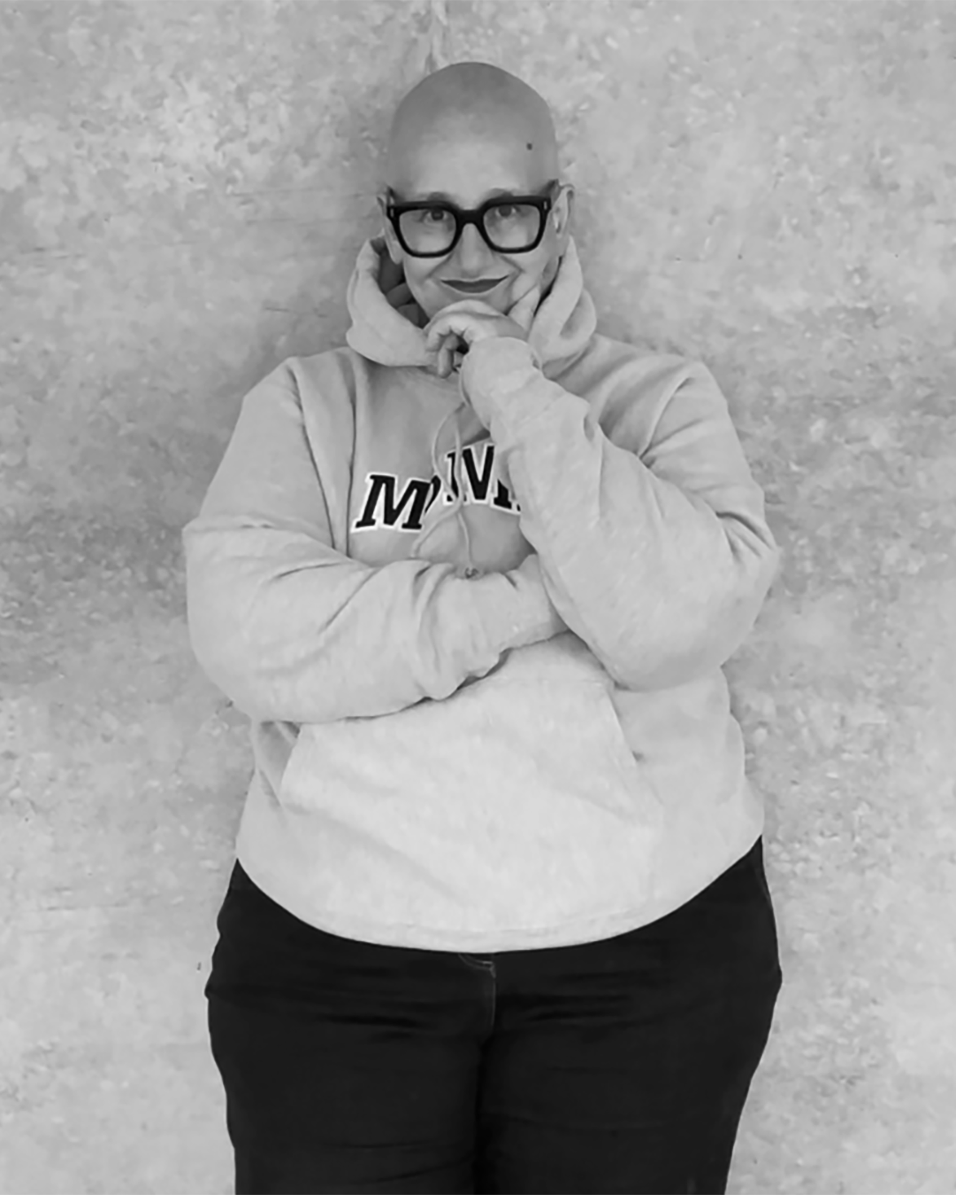 A woman with a shaved head and glasses standing against a plain background, wearing a hoodie with her arms crossed, smiling softly.