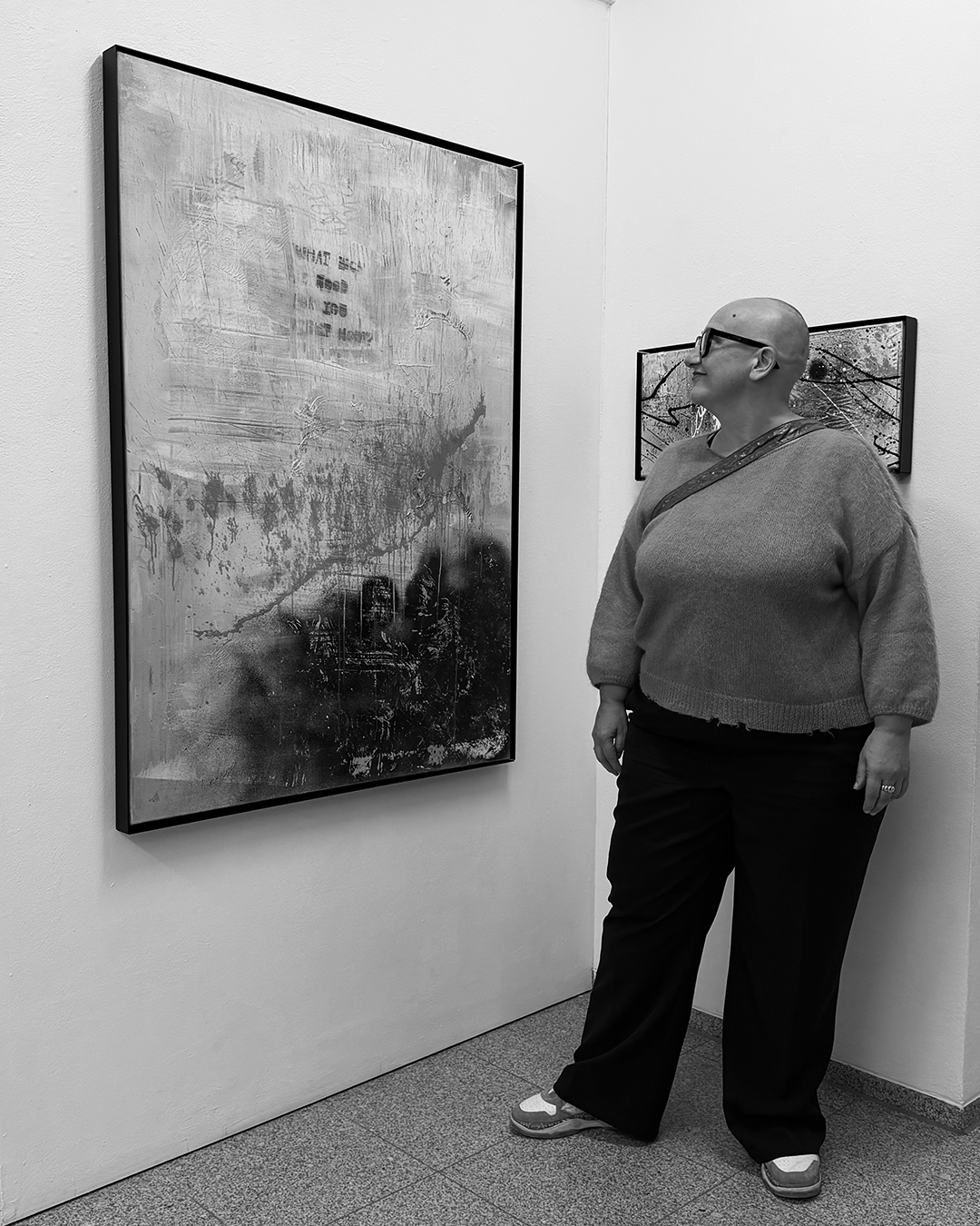 A woman with glasses and a shaved head standing in an art gallery, observing a large abstract painting on the wall.