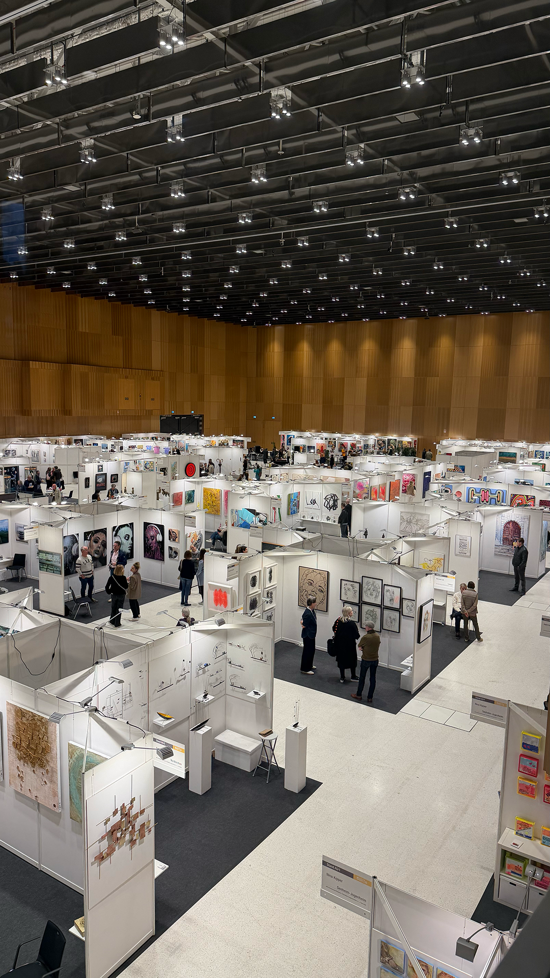 Art exhibition in a large indoor gallery with white partition walls displaying paintings and sculptures, visitors walking and viewing artworks.