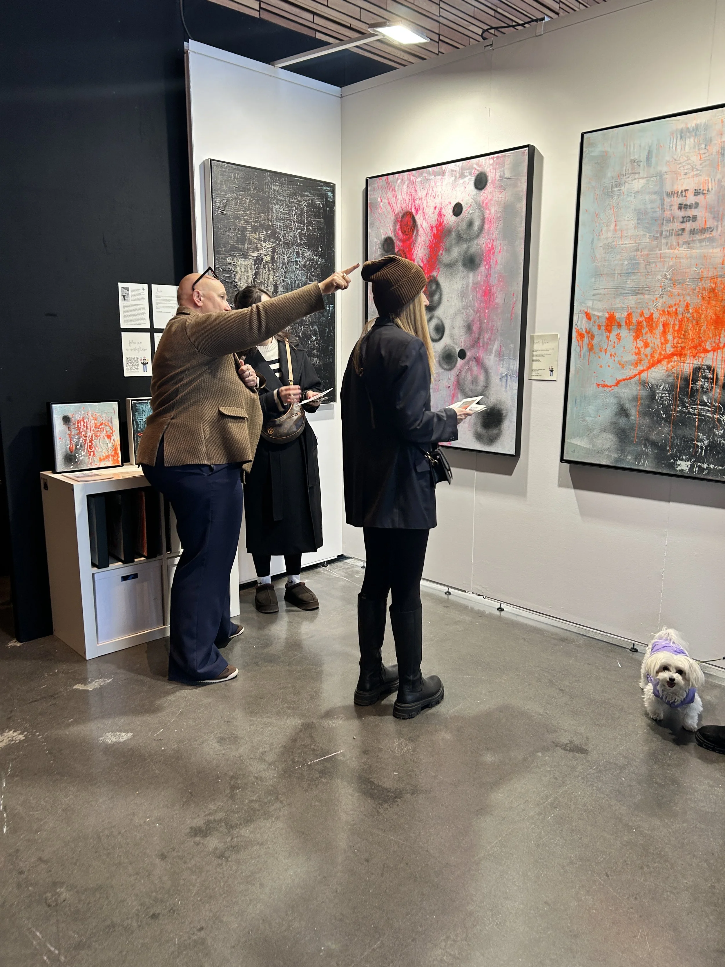Three people and a small dog viewing abstract art paintings in an art gallery. The man is pointing at one of the artworks, while two women look at the paintings and a small dog is on the floor near them.