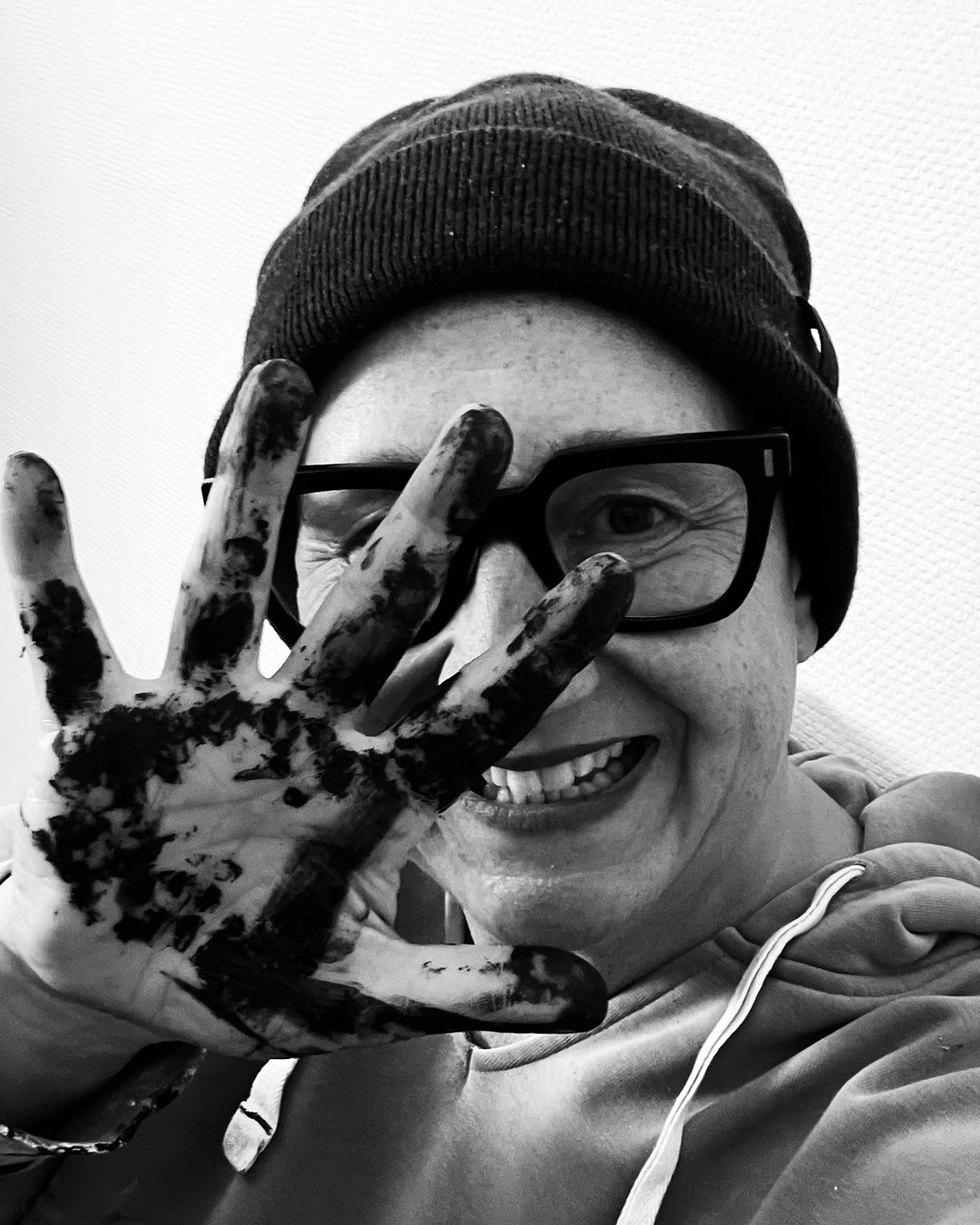 A smiling person wearing glasses, a beanie, and a hoodie, holding their hand up to their face covered in black paint, with a textured wall in the background.