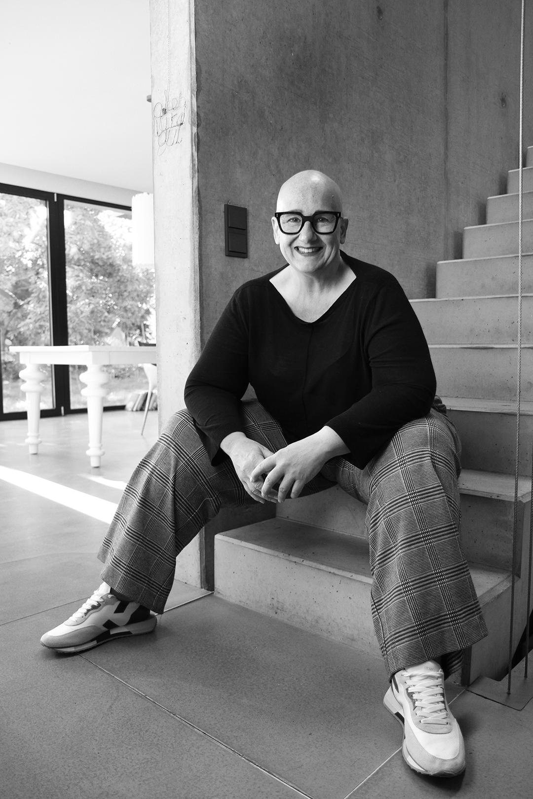A smiling, bald woman with glasses sitting on stairs in a modern interior with large windows and a concrete wall.