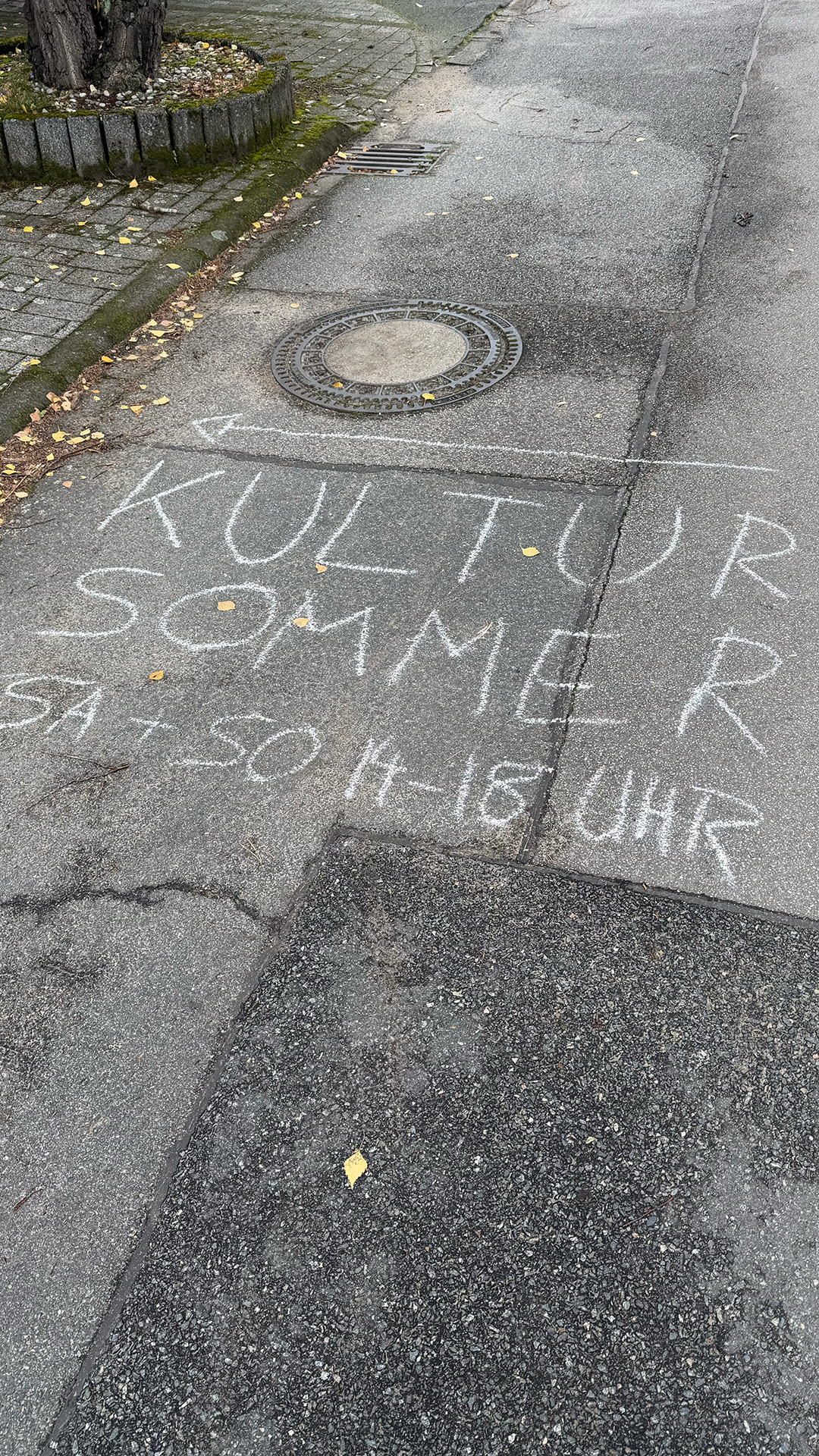 Chalk writing on a sidewalk that says, 'KULTUR SOMMER 20-24' near a tree.