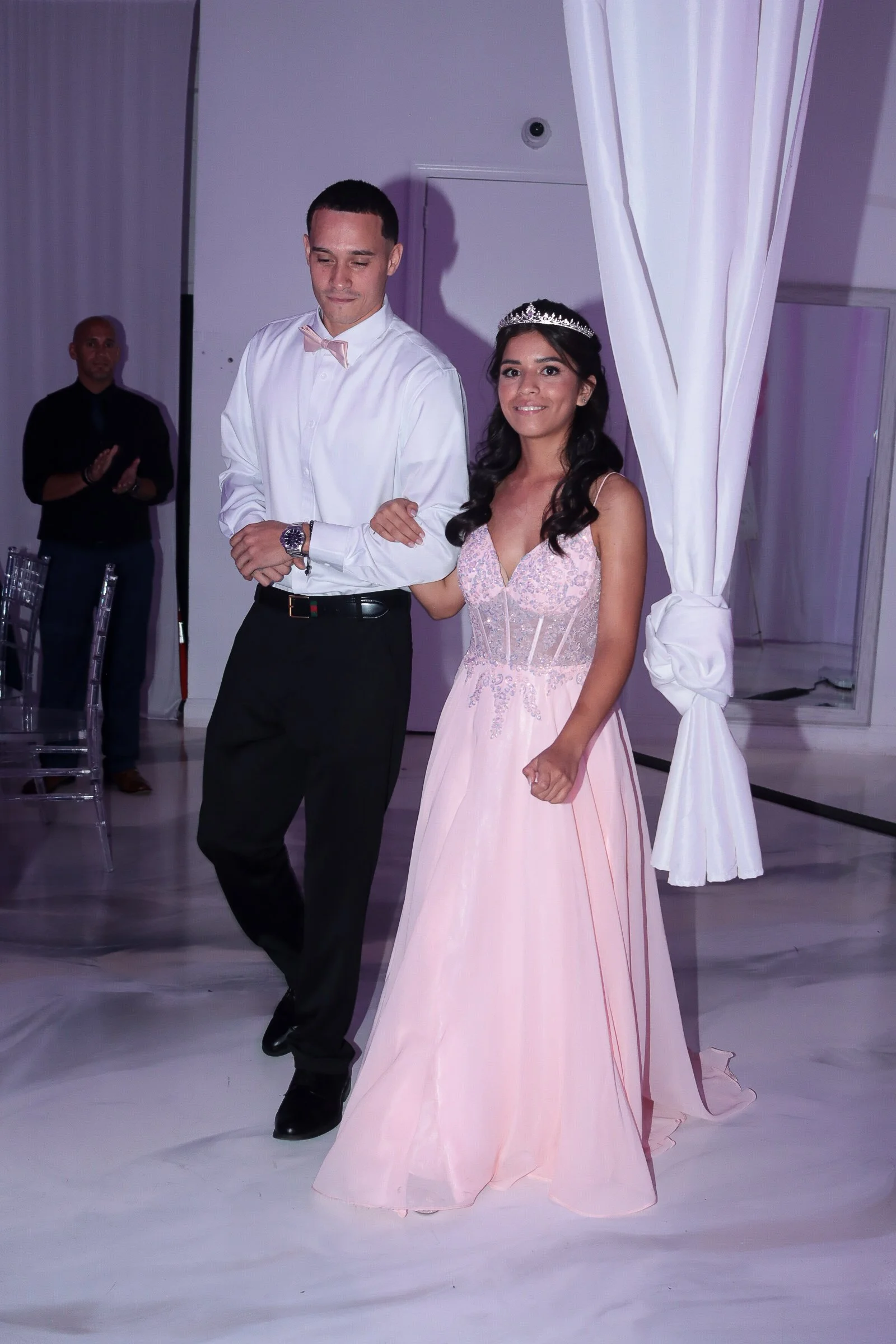 A young couple at a formal event, the woman wearing a strapless pink gown and a tiara, and the man dressed in a white shirt with a bow tie, holding hands and walking together in an elegant setting.