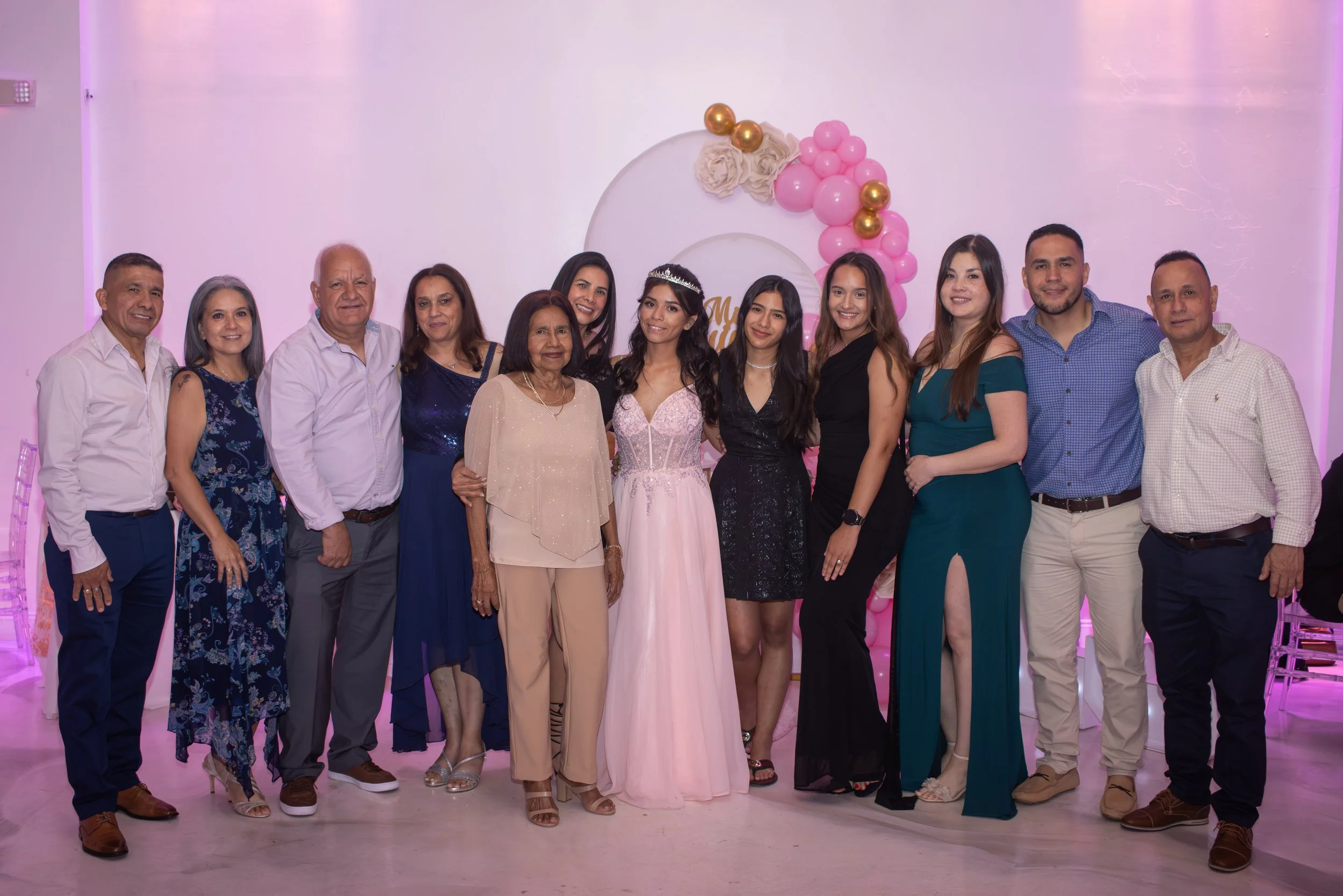 Family and friends gathered for a birthday celebration, with a young woman wearing a pink dress and tiara, posing in the center with other adults around her, behind pink and gold balloons and decorative flowers.