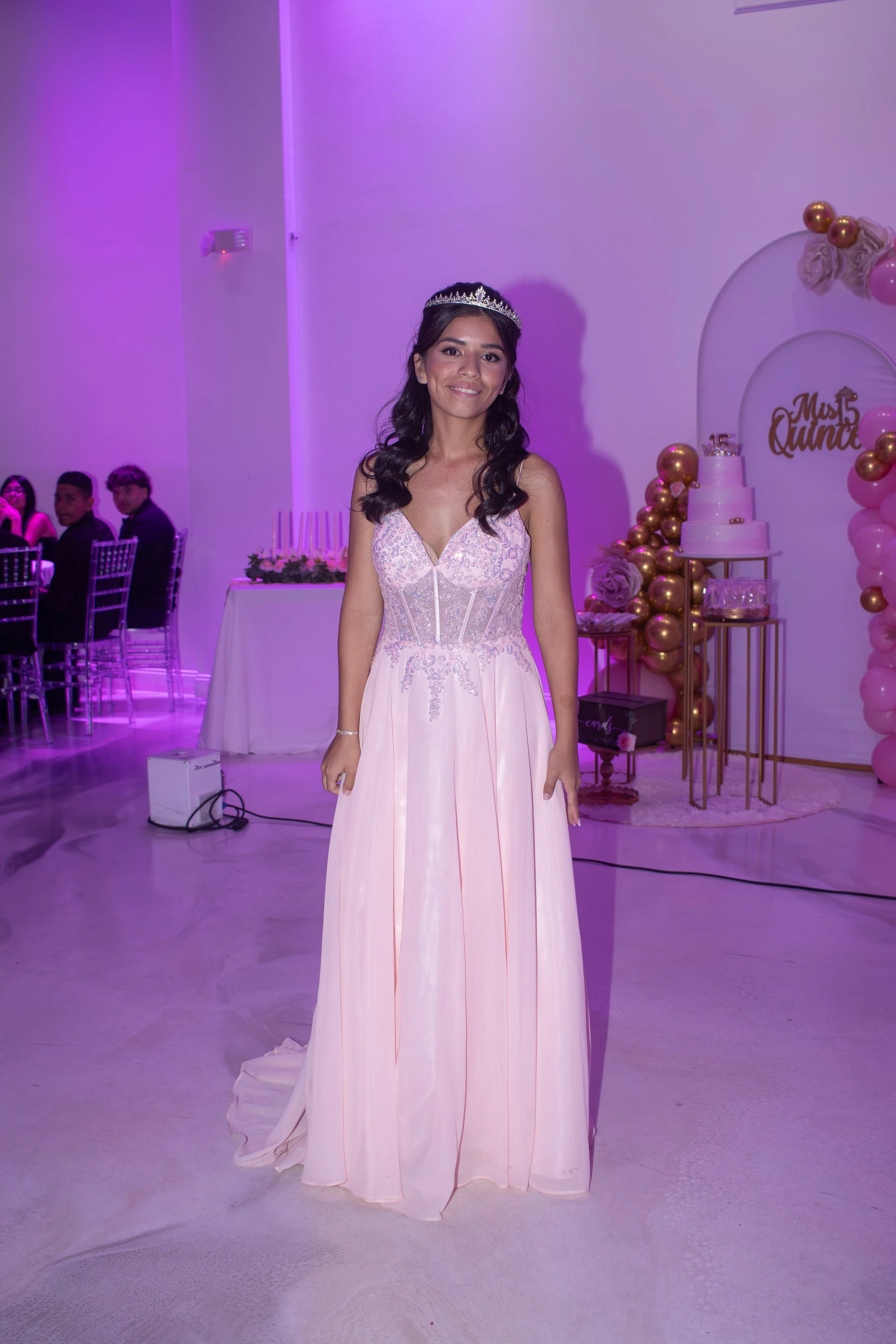 A young woman in a pink dress with a crown standing in a decorated event space for a quinceañera celebration.