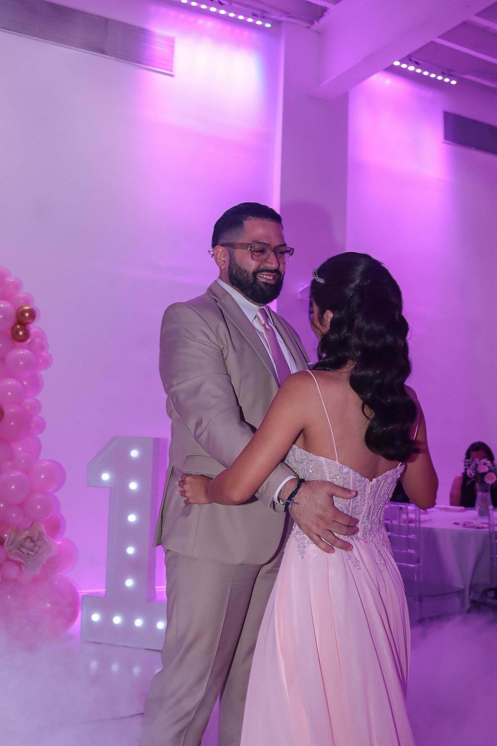 A man and woman dance closely at a celebration, surrounded by pink and purple lighting, with decorative balloons and illuminated letters in the background.