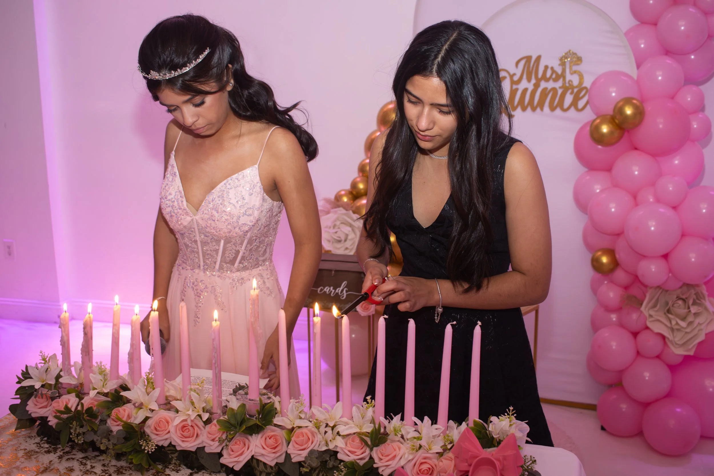 Two women at a birthday celebration, one in a pink dress with a tiara, and the other in a black dress, lighting candles on a cake decorated with pink roses and surrounded by pink and gold balloons.