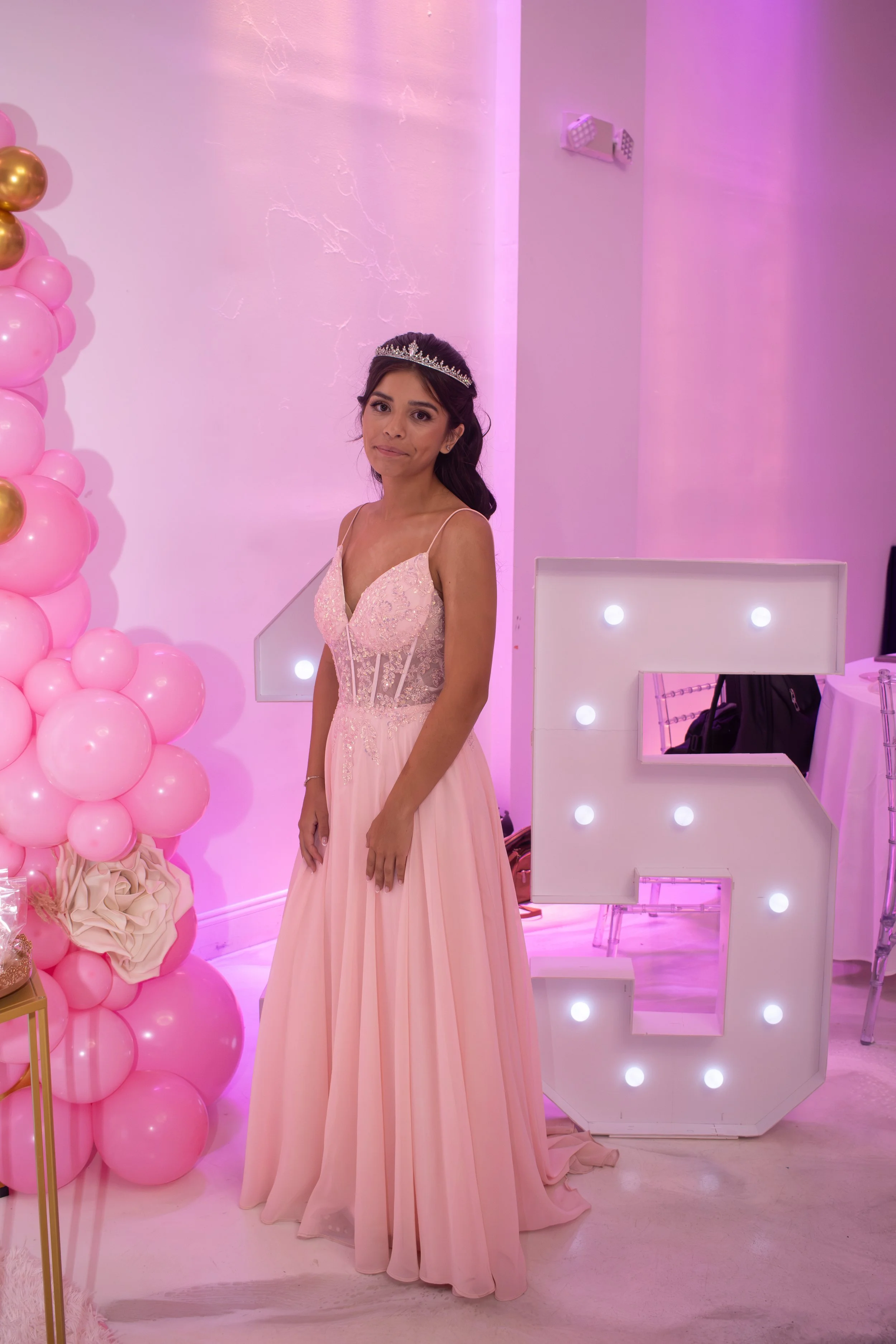 Young woman wearing a pink dress and tiara standing in front of pink and gold balloon decoration, with a lit letter Wonder sign and mirrored vanity in the background.