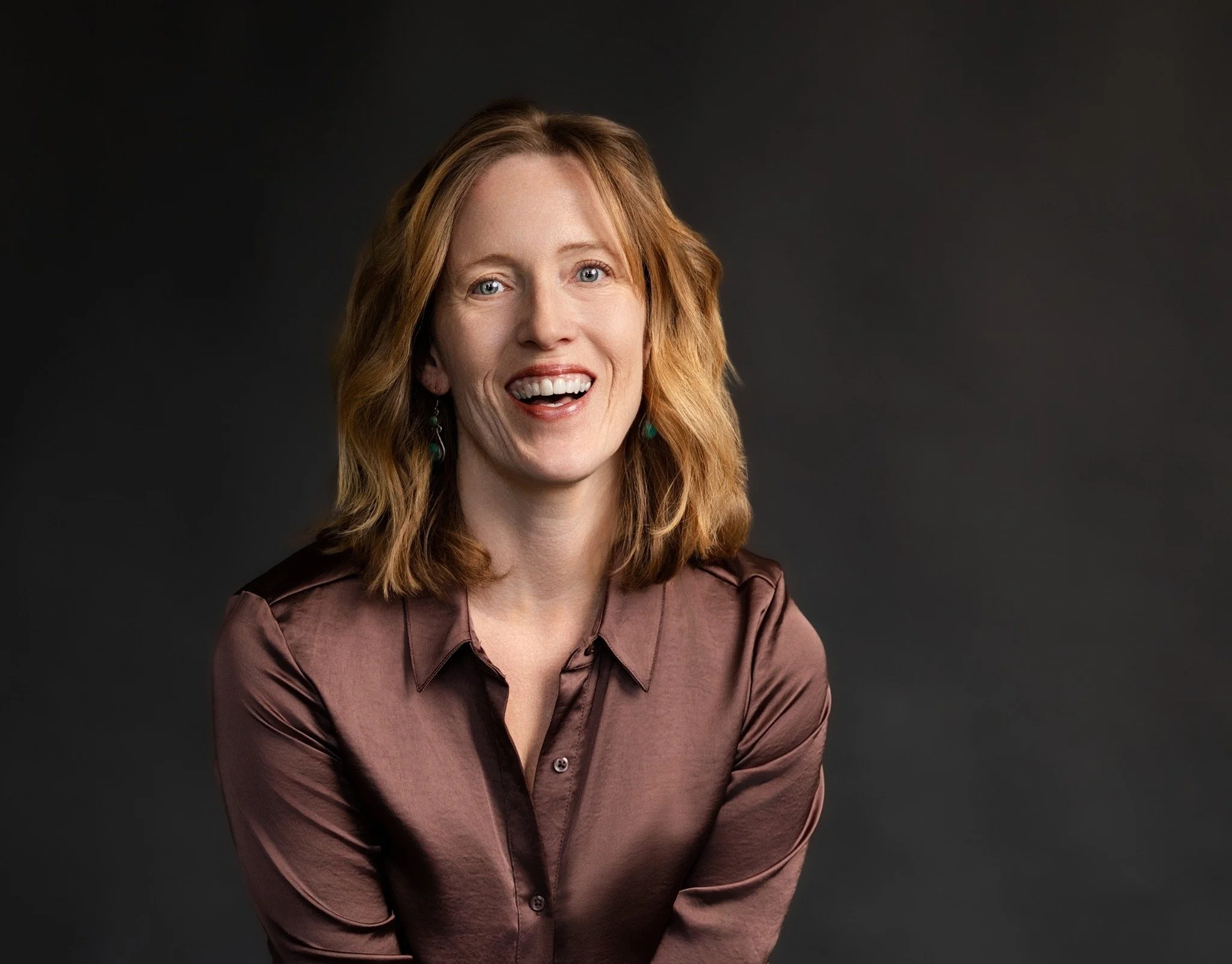 Jen Knox. An author with long red hair, blue eyes, and a wide smile, wearing a brown satin button-up shirt, against a dark grey background.