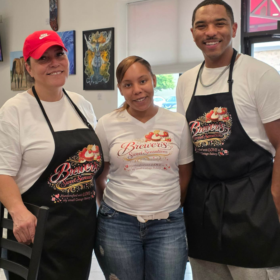 Three people standing inside a bakery, wearing bakery aprons and smiling at the camera. The woman on the left is wearing a red Nike cap, white t-shirt, and black apron. The woman in the middle is wearing a white t-shirt and jeans. The man on the right is wearing a white t-shirt and black apron. Artwork hangs on the wall behind them, and a window shows the outside.