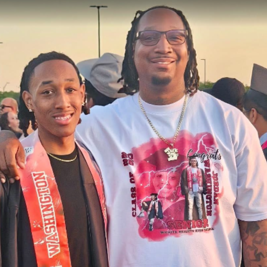 Two men smiling at an outdoor graduation celebration, one young man with a graduation stole and the other in a T-shirt with graduation-themed graphics.