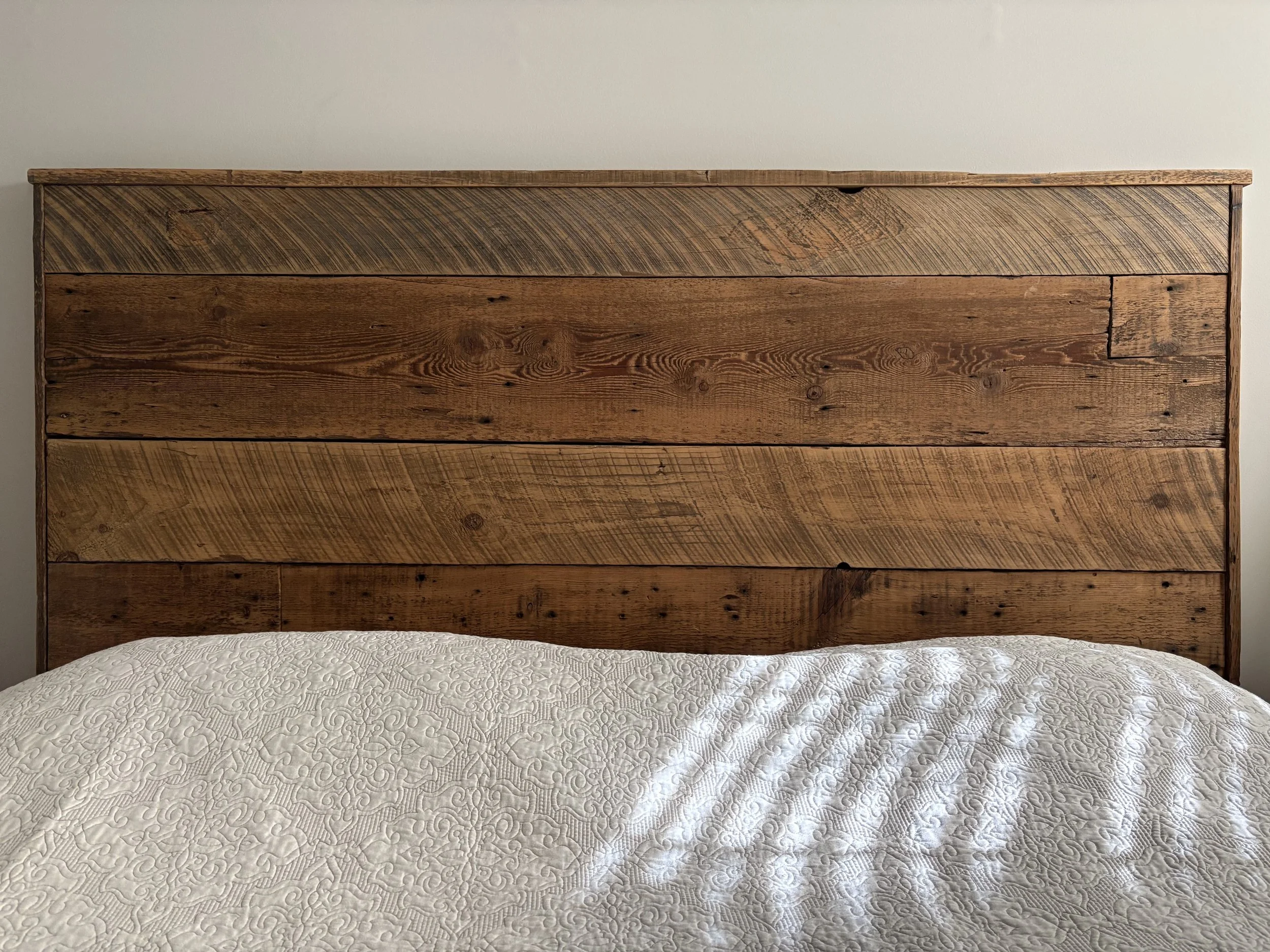 Close-up view of a bed with a quilted cream-colored bedspread and a headboard made of rustic, stained wood planks.