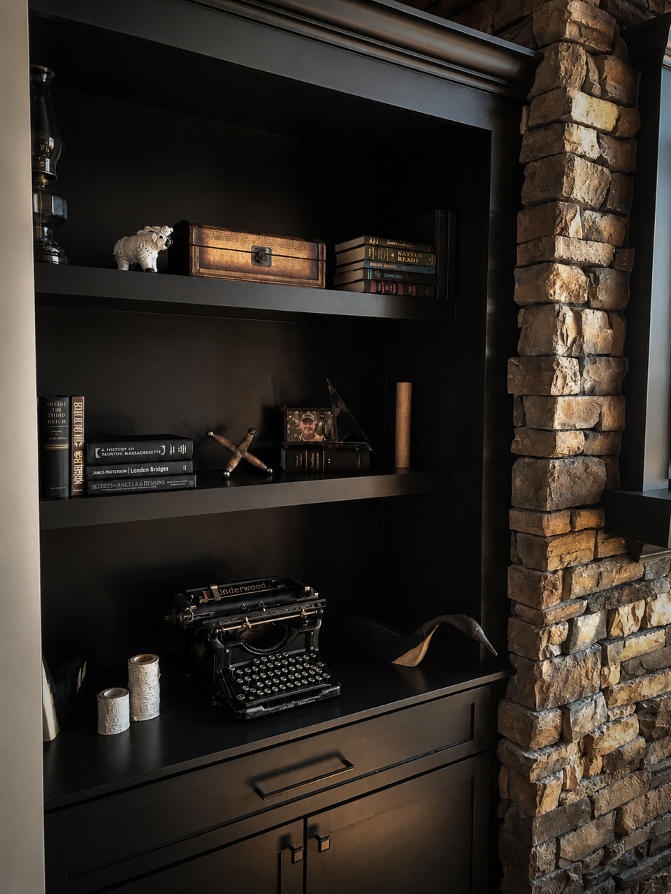 A black bookshelf with decorative items, books, an old typewriter, and a small sculpture of a bird, next to a stone wall.