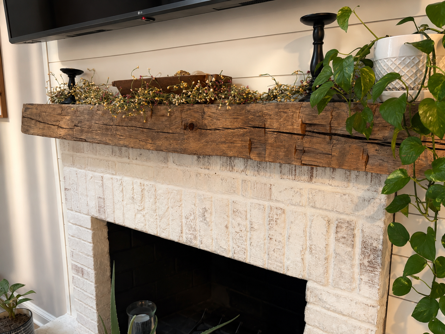 A fireplace with a white brick surround and a rustic wooden mantel decorated with plants and black candlesticks.