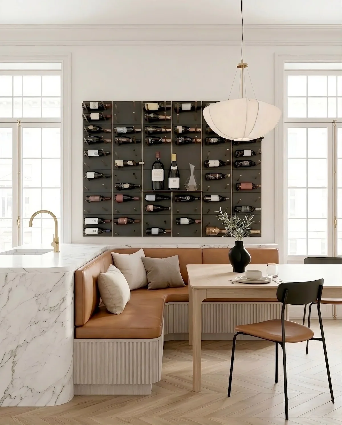 A modern dining room with a wine rack on the wall, a table set with plates, glasses, and a vase with greenery, a leather banquette with pillows, and large windows in the background.