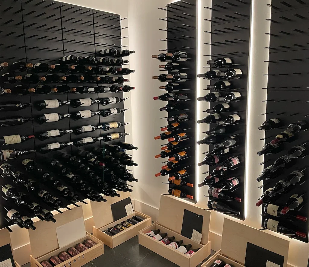 A modern wine cellar with black wall-mounted racks holding numerous bottles of wine, illuminated by vertical light strips, with wooden crates of wine bottles on the floor.