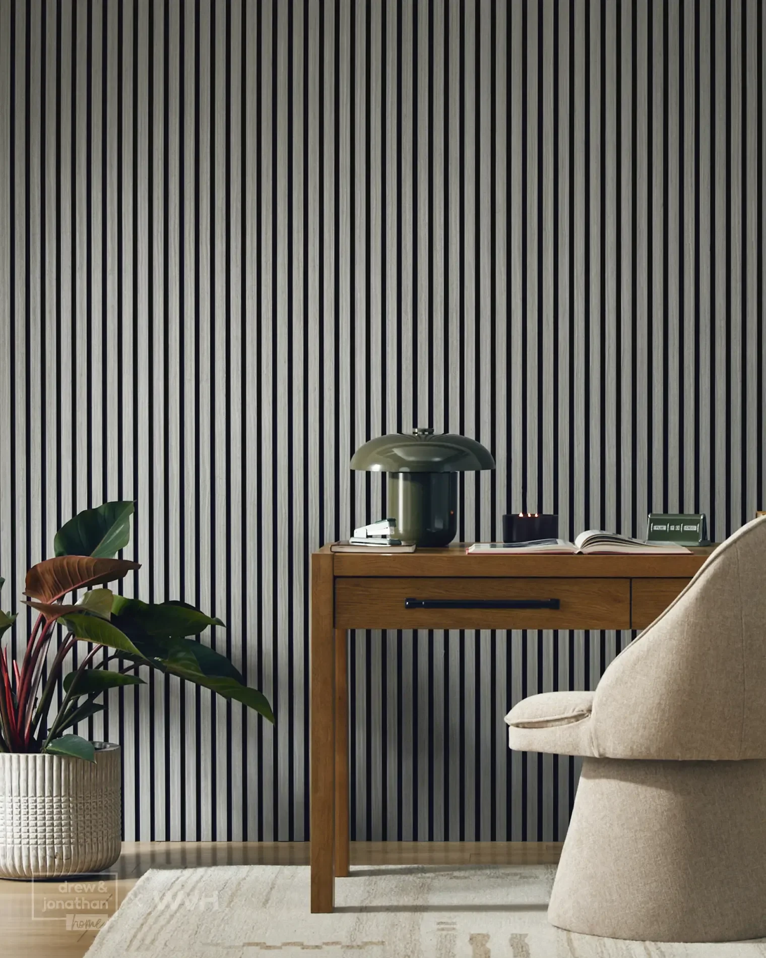 Modern home office with vertical gray striped wall, wooden desk, beige upholstered armchair, decorative table lamp, open books, and potted houseplant with large green leaves.