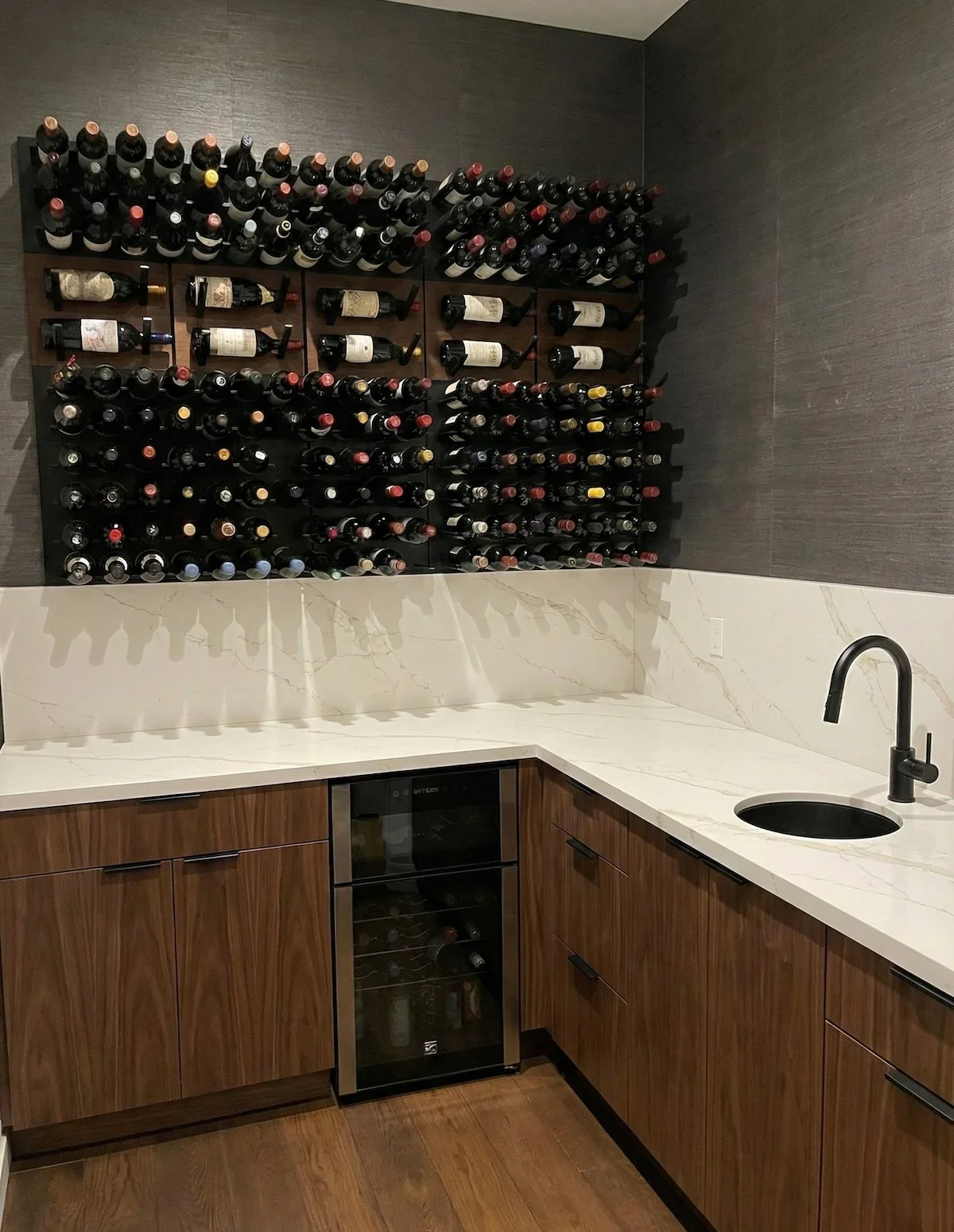 A wine storage rack filled with wine bottles, mounted on a dark wall above a white marble countertop in a modern kitchen.
