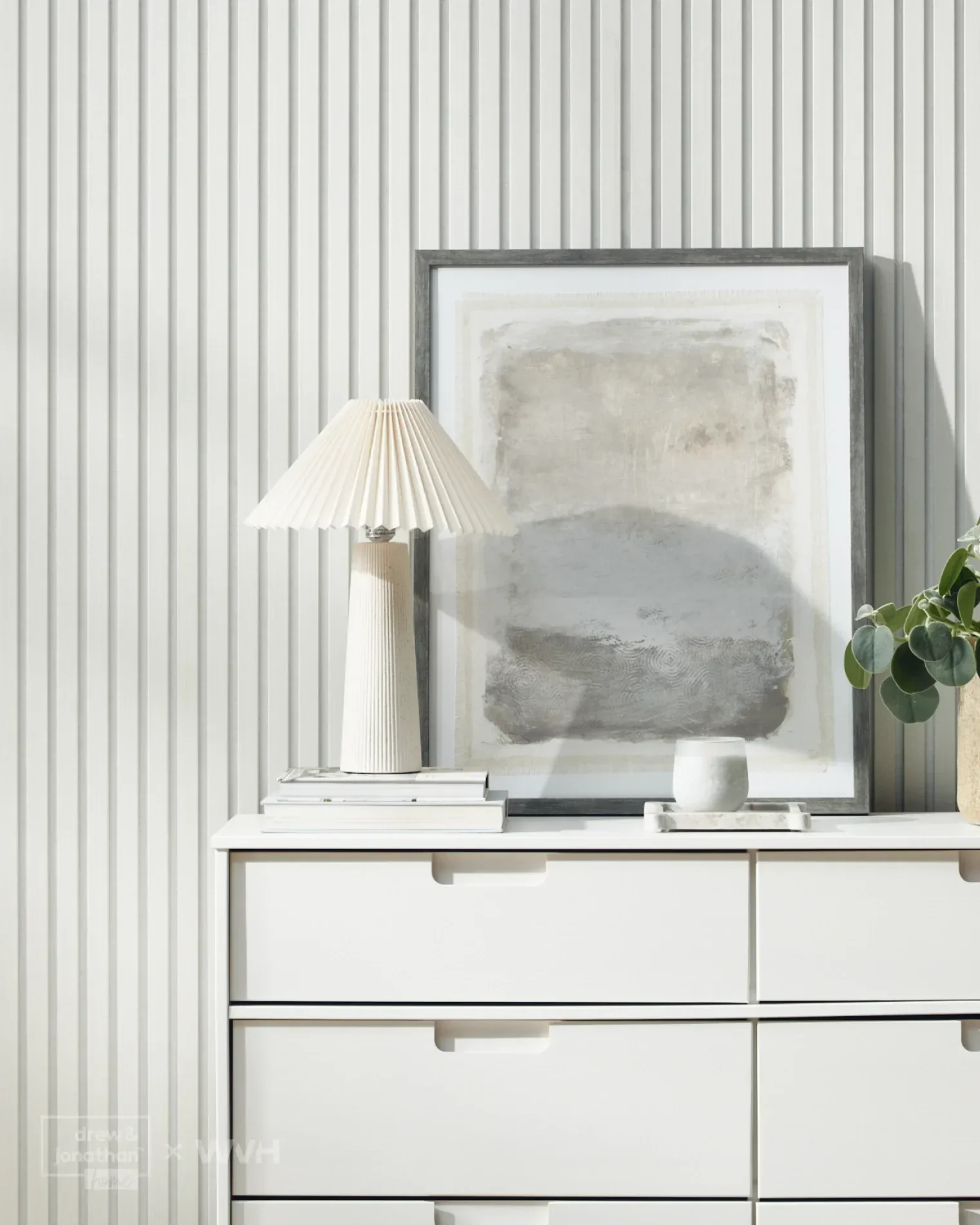 A white sideboard with three drawers, a table lamp, a stack of books, an empty white bowl, a framed abstract artwork, and a potted plant on top against a vertically ridged white wall.