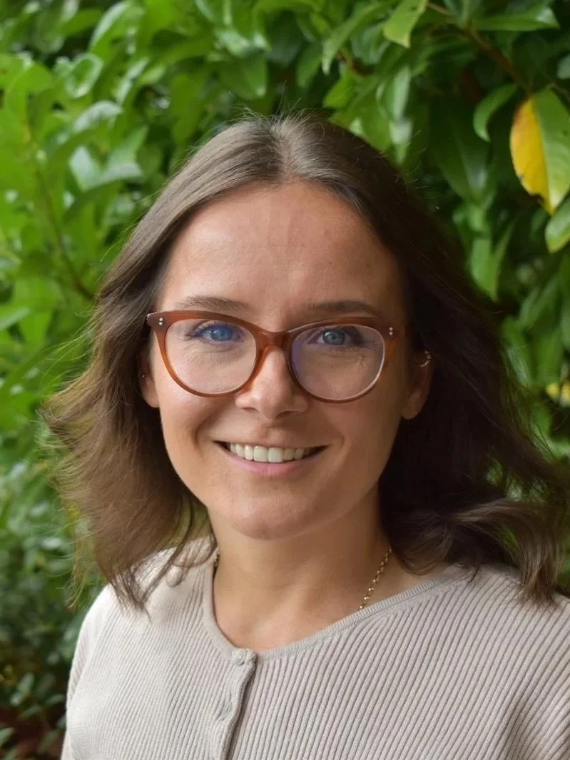 A woman with shoulder-length brown hair, wearing glasses and a beige top, standing outdoors with green foliage in the background.