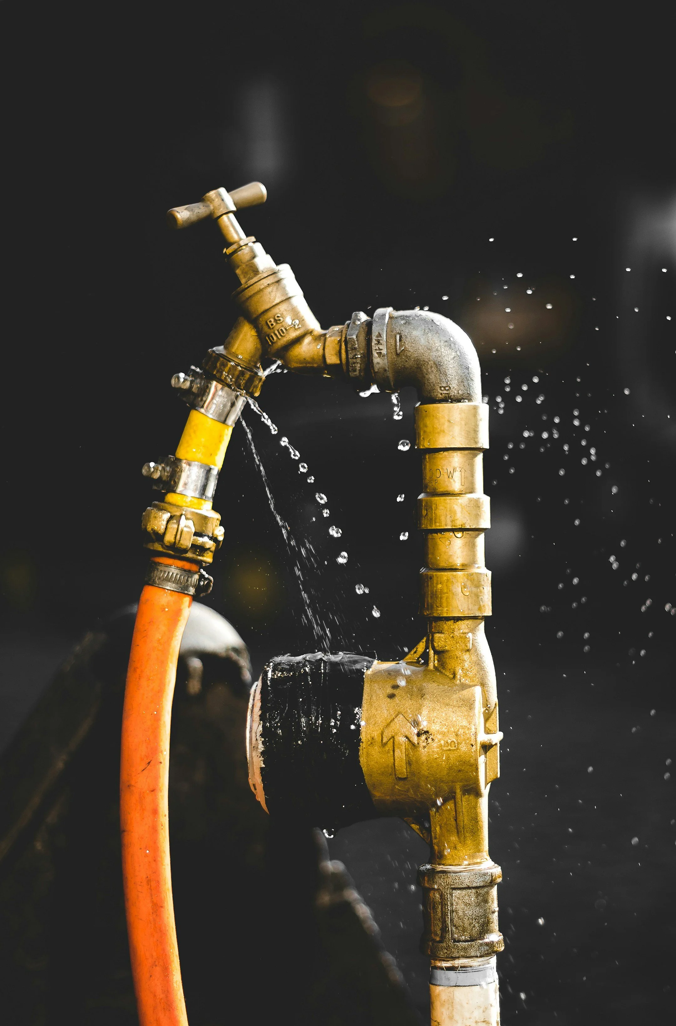 Un robinet en laiton coulant de l'eau, connecté à des tuyaux en plastique orange et jaune, avec de l'eau qui s'écoule et éclabousse sur un fond sombre.