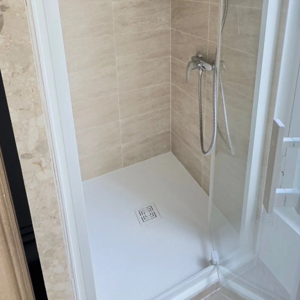 une cabine de douche avec des murs en carrelage beige et un sol blanc, équipée d'une douche à main et d'une ridelle en verre