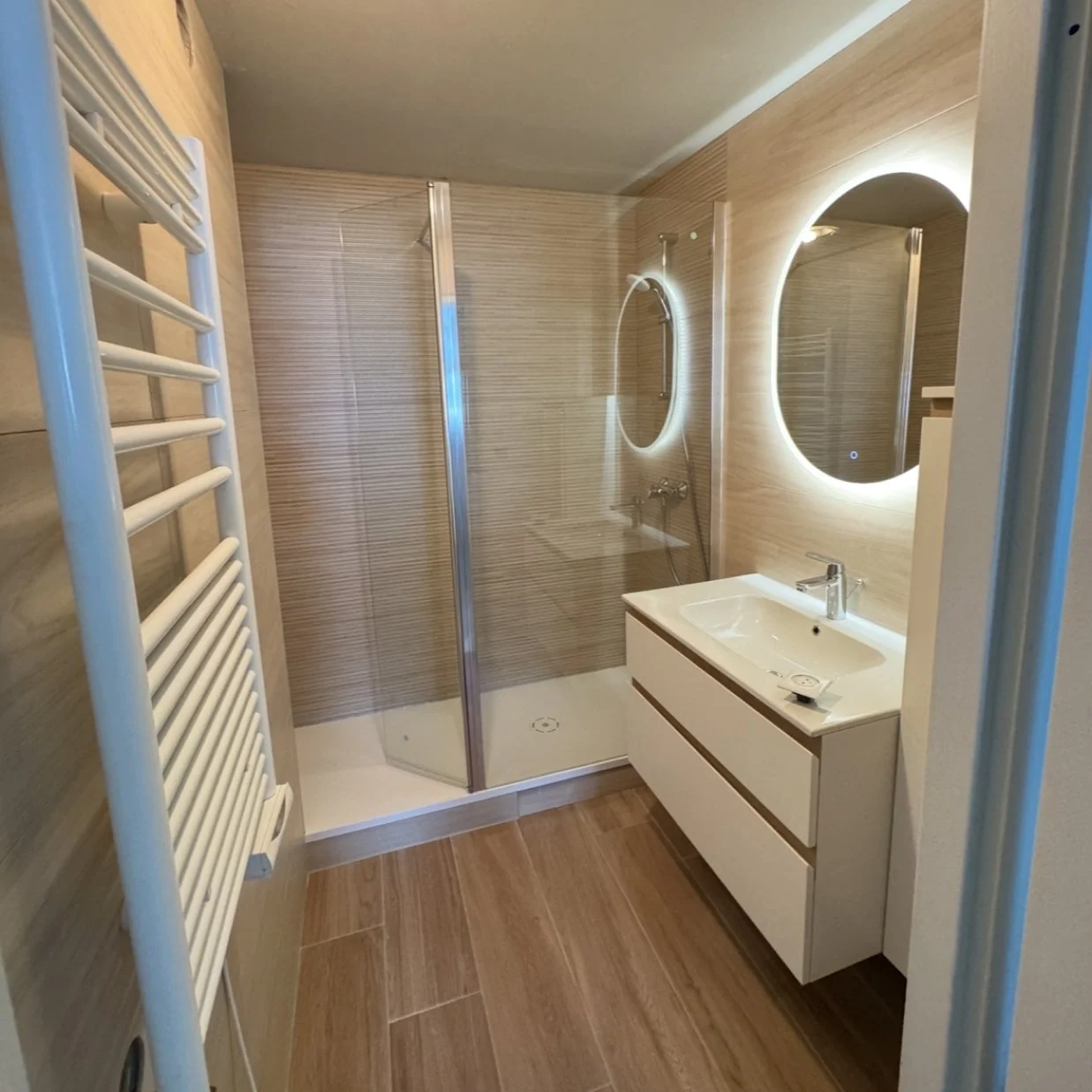 Salle de bain moderne avec douche à l'italienne, miroir rond avec éclairage LED, lavabo blanc avec meuble, et radiateur sèche-serviettes blanc.